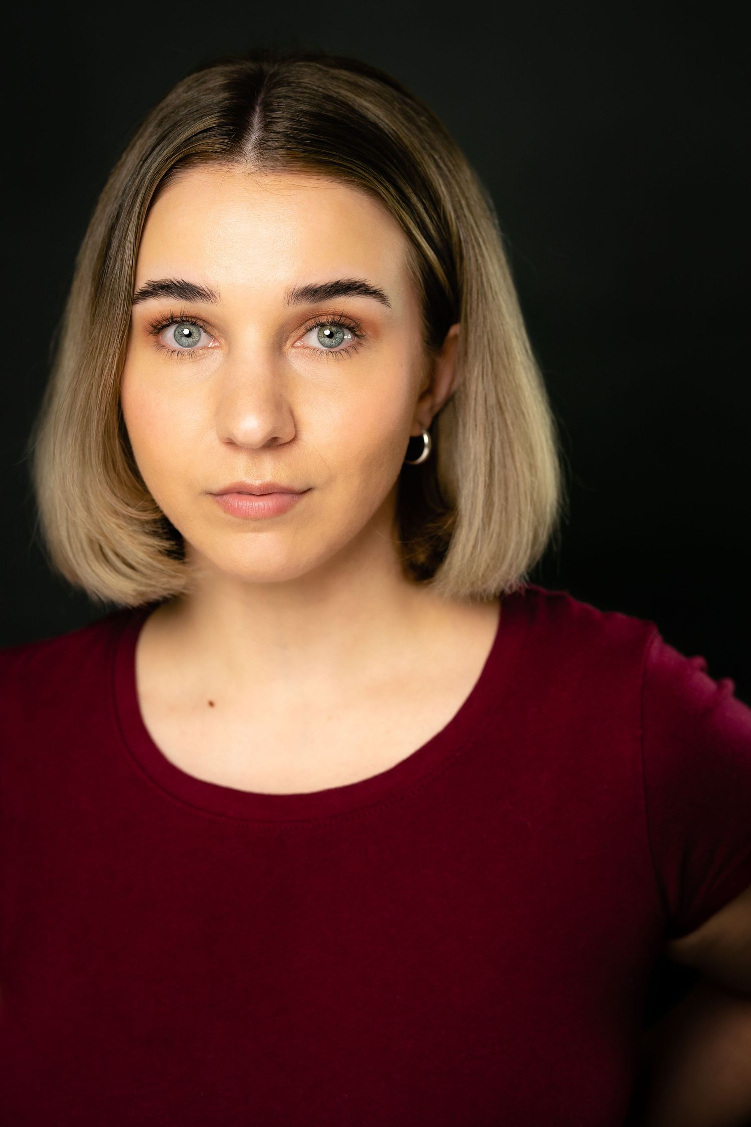 Young woman with blonde bob hair and blue eyes wearing a red top against a dark background.
