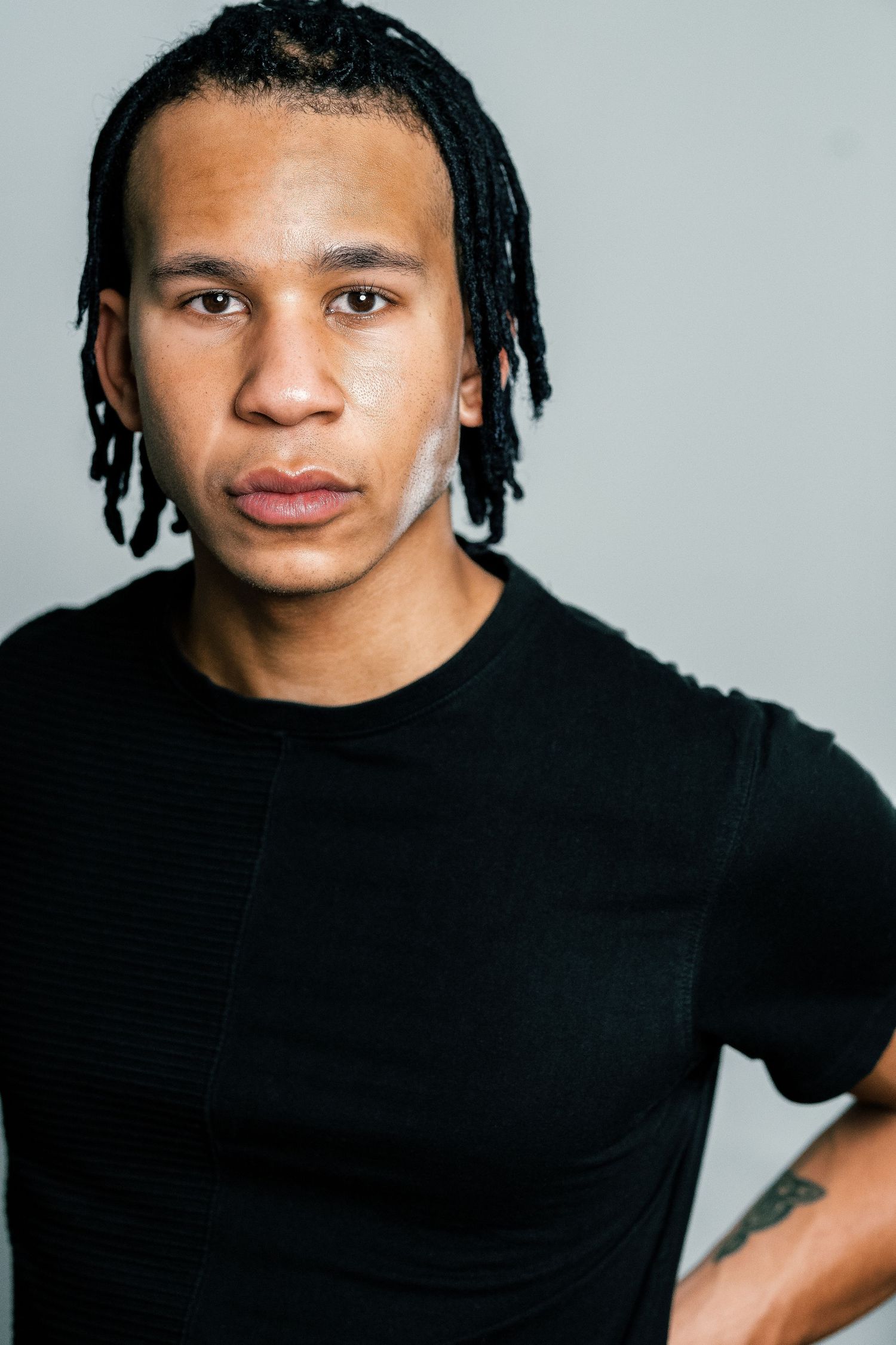 Young Black man with dreadlocks wearing a black fitted t-shirt, posing against a gray background.