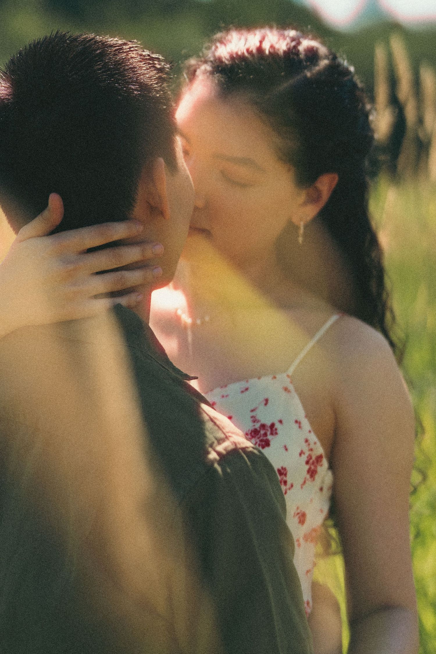 Couple sharing a romantic kiss outdoors in a sunlit green field, woman in floral dress with hand on mans neck.