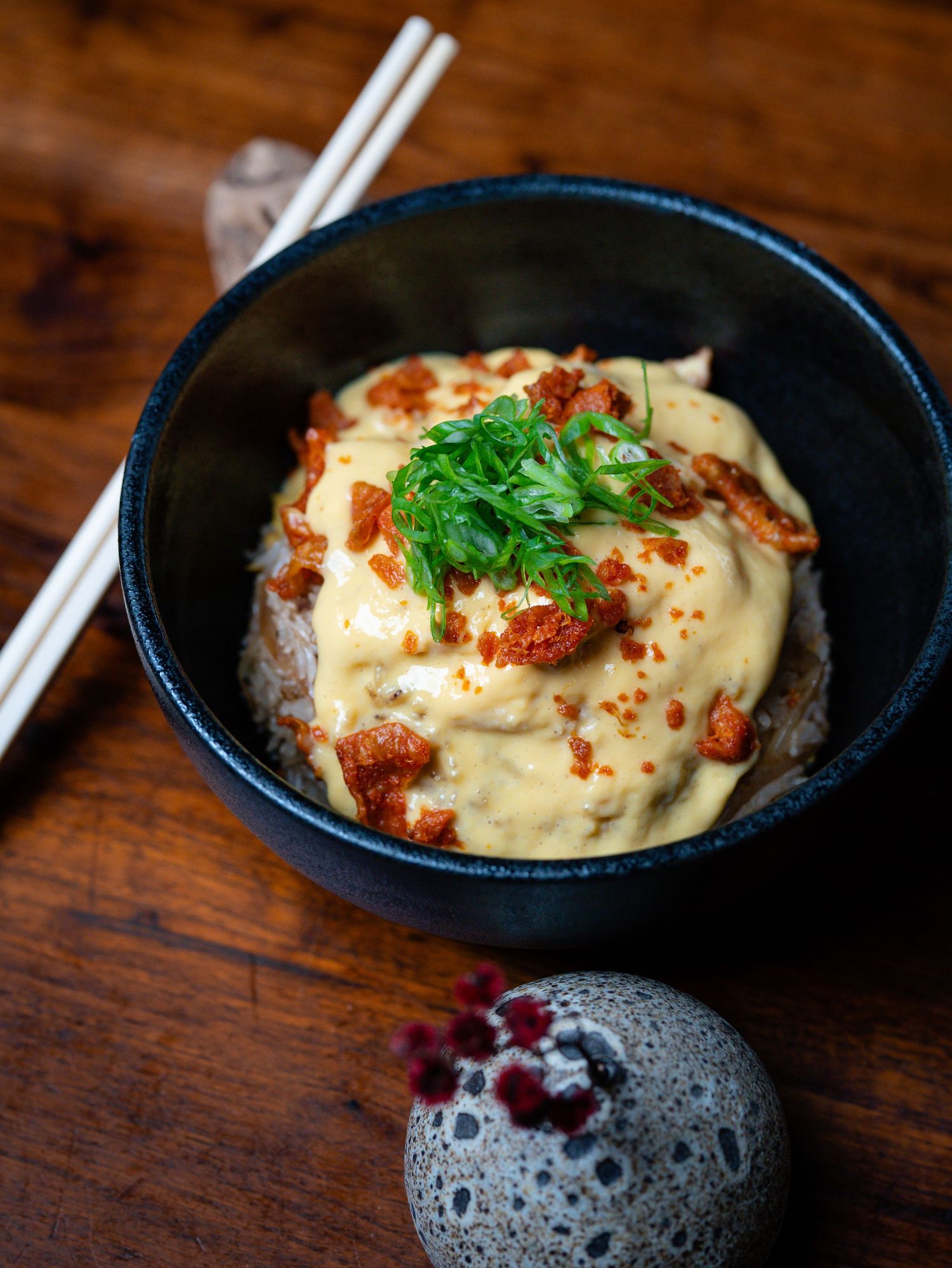 A dark ceramic bowl of rice topped with creamy sauce, crispy garnish, and fresh herbs, with chopsticks on a wooden table.