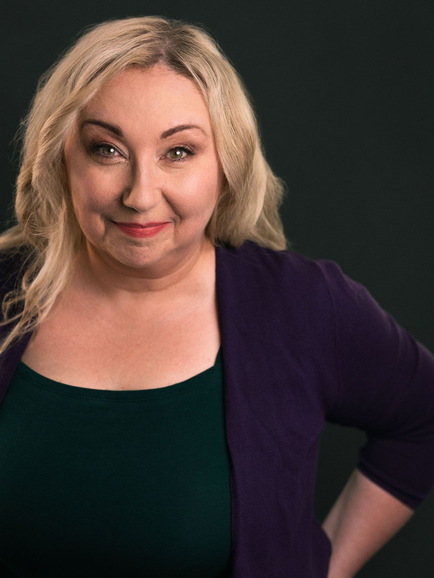 Blonde woman in green top and purple cardigan smiling confidently against dark background.