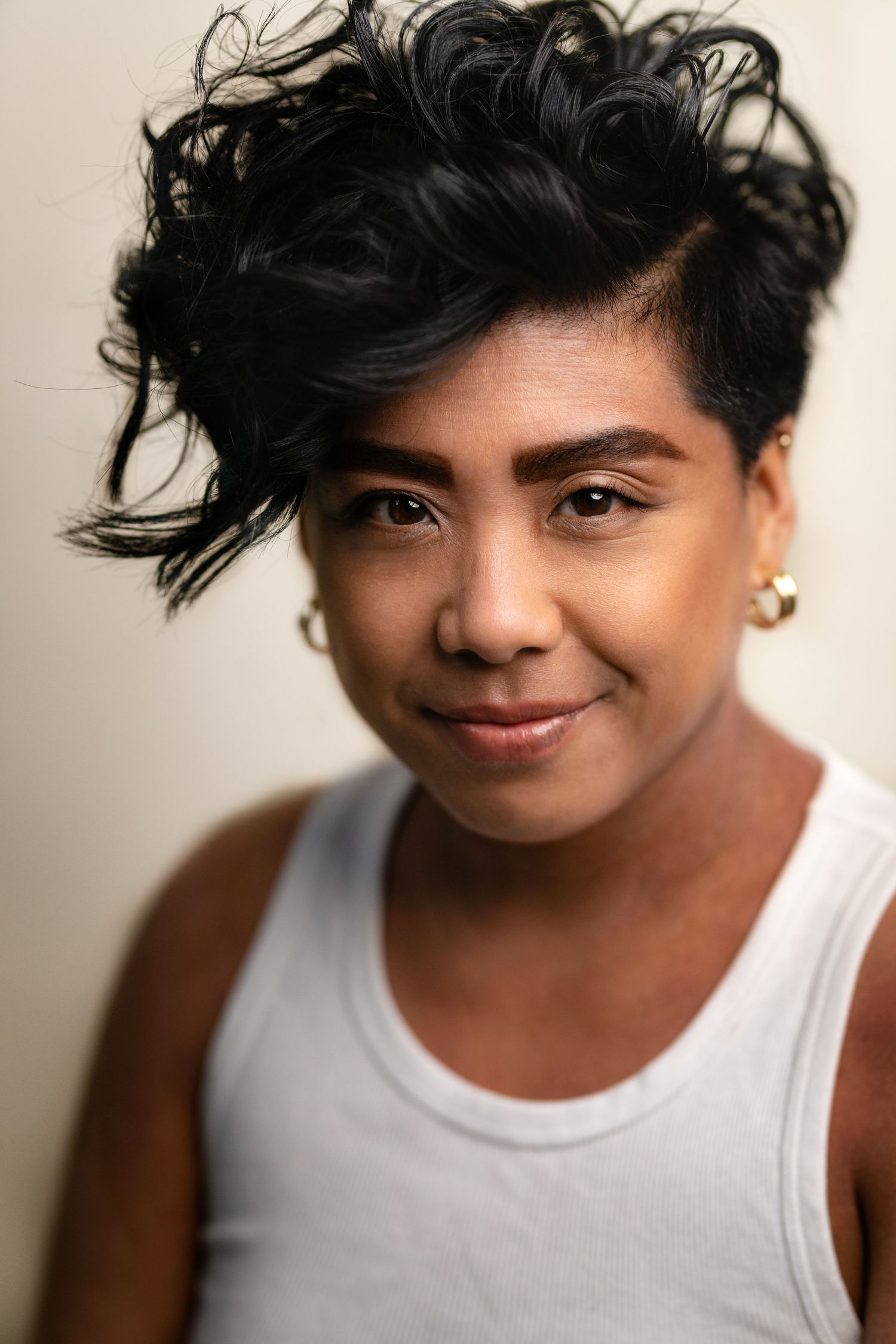Young person with short curly black hair and gold hoop earrings wearing a white tank top, smiling softly.