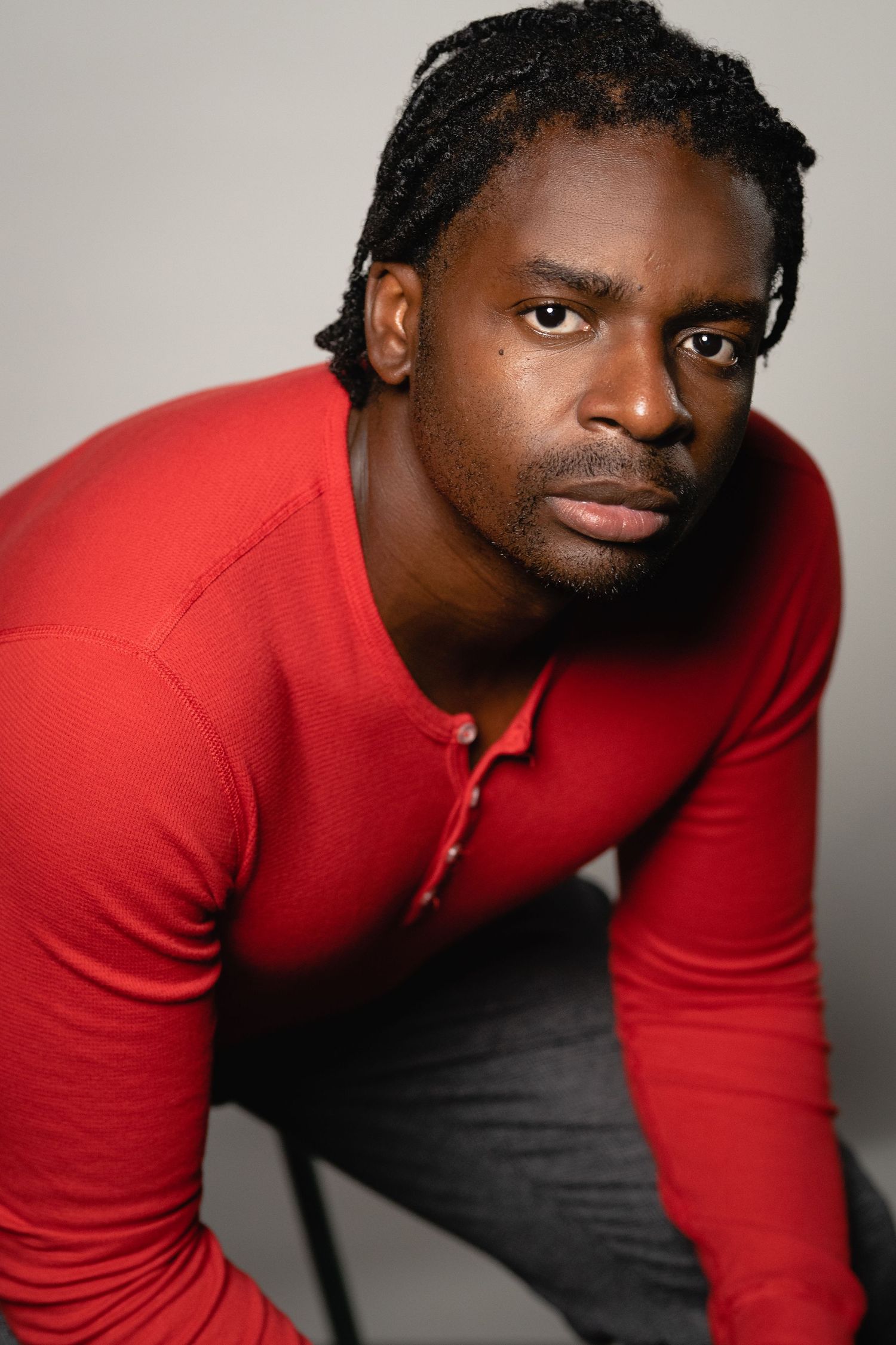 A man with braided hair wearing a red long-sleeve henley shirt poses seriously against a gray background.
