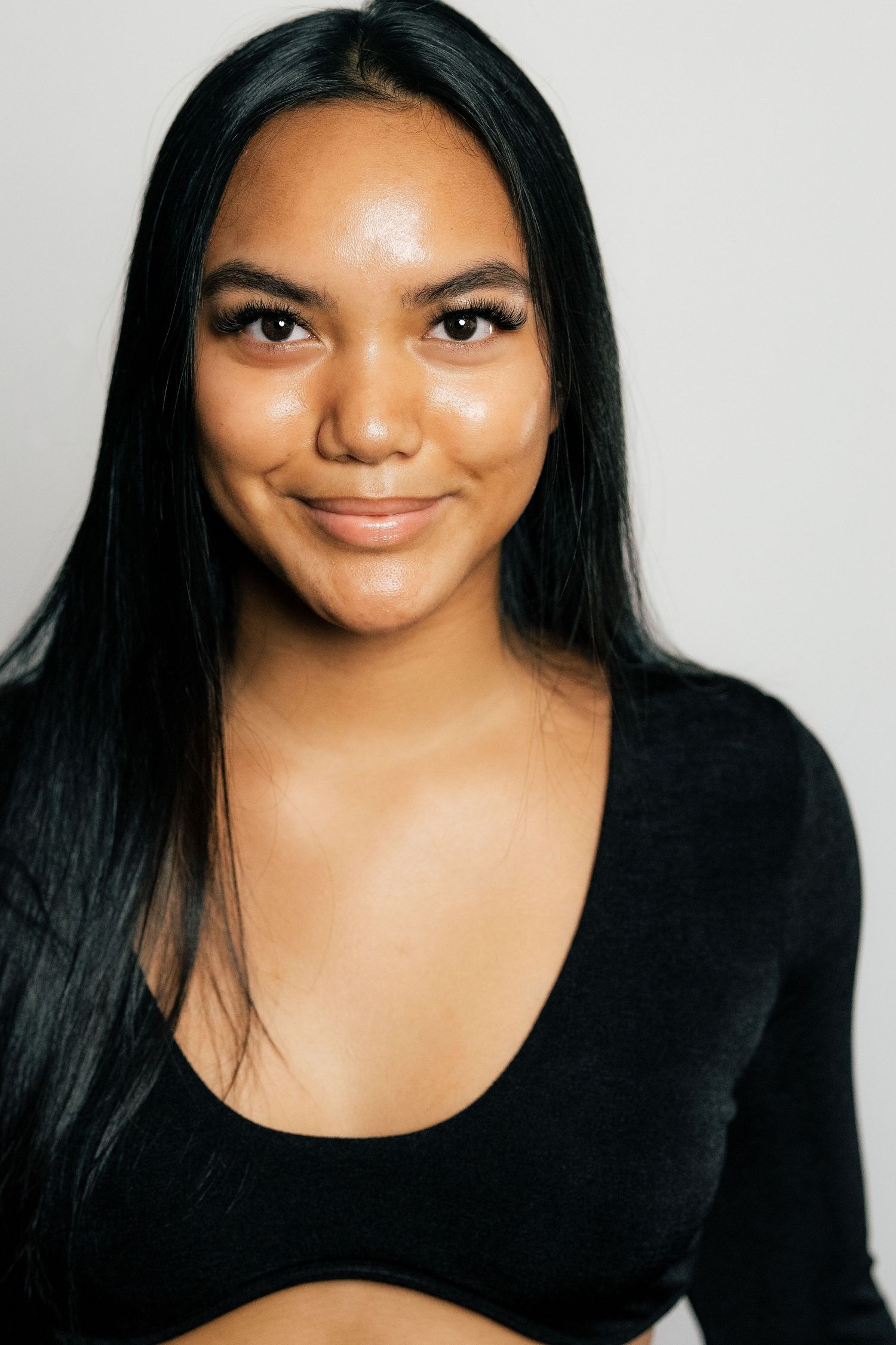 Young woman with long black hair smiling, wearing a black crop top against a light gray background.