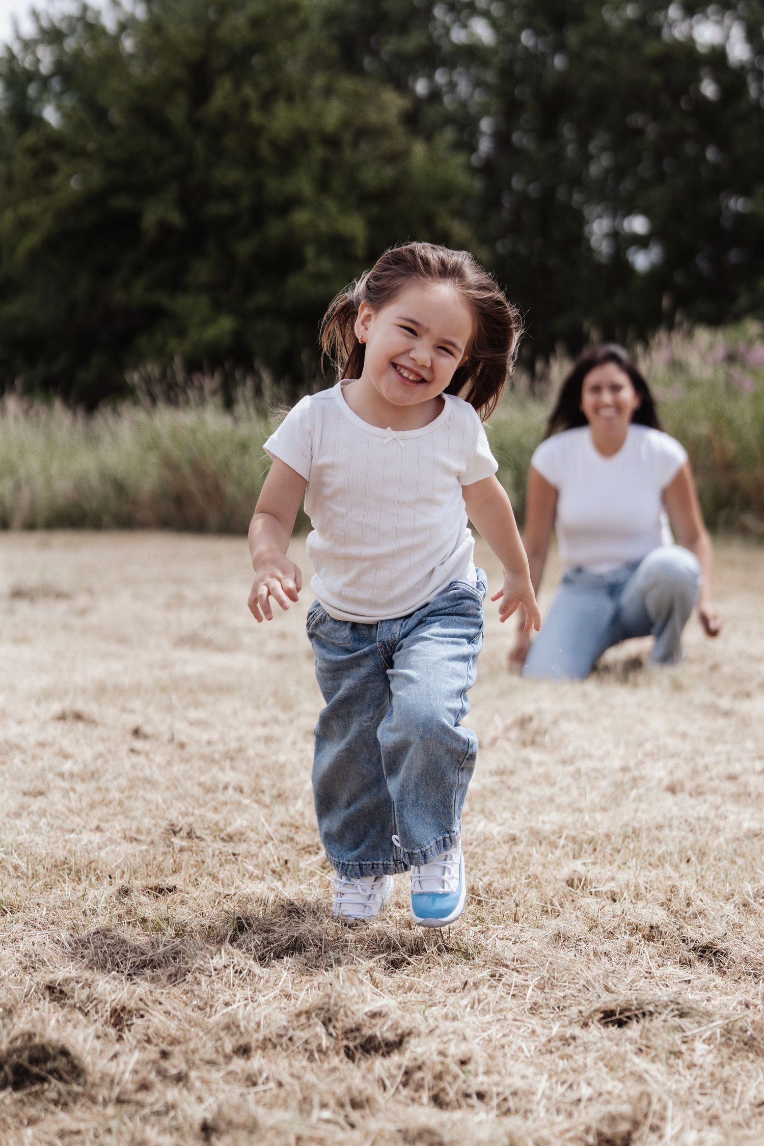 A joyful little girl with pigtails runs through a field while her mother kneels smiling in the background.