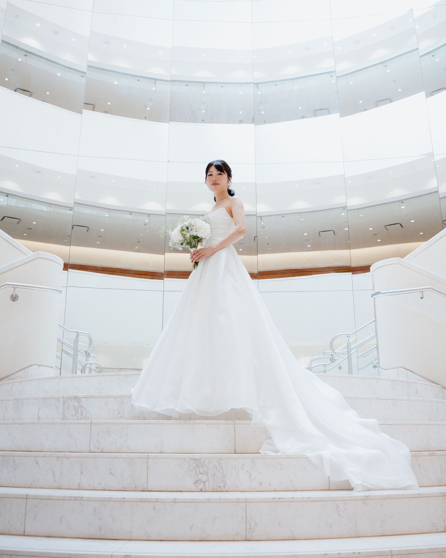 Bride in a flowing white wedding gown holding a bouquet, standing on marble stairs in an elegant circular venue.