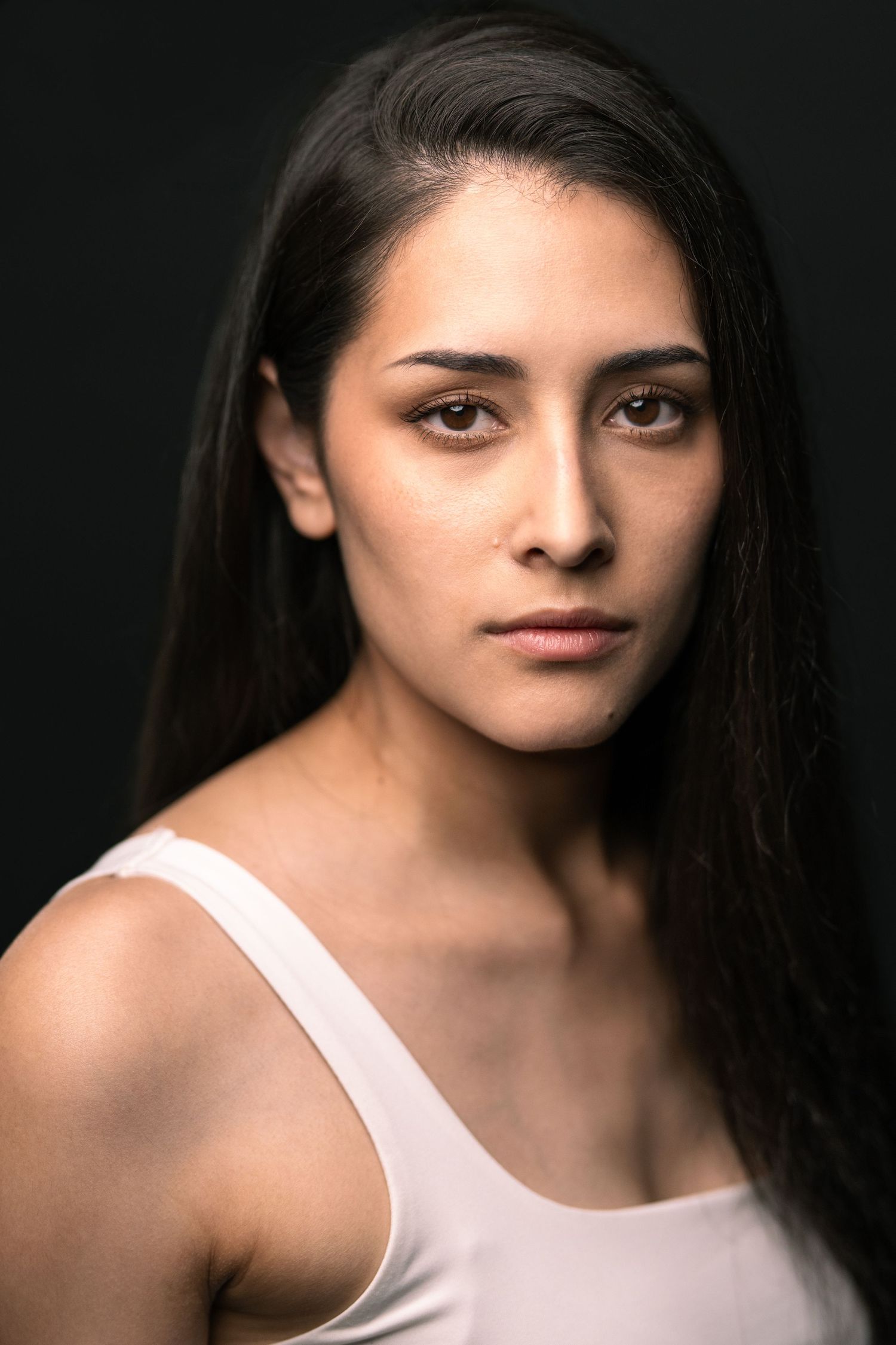 A young woman with long dark hair wearing a white tank top poses against a dark background with a serious expression.