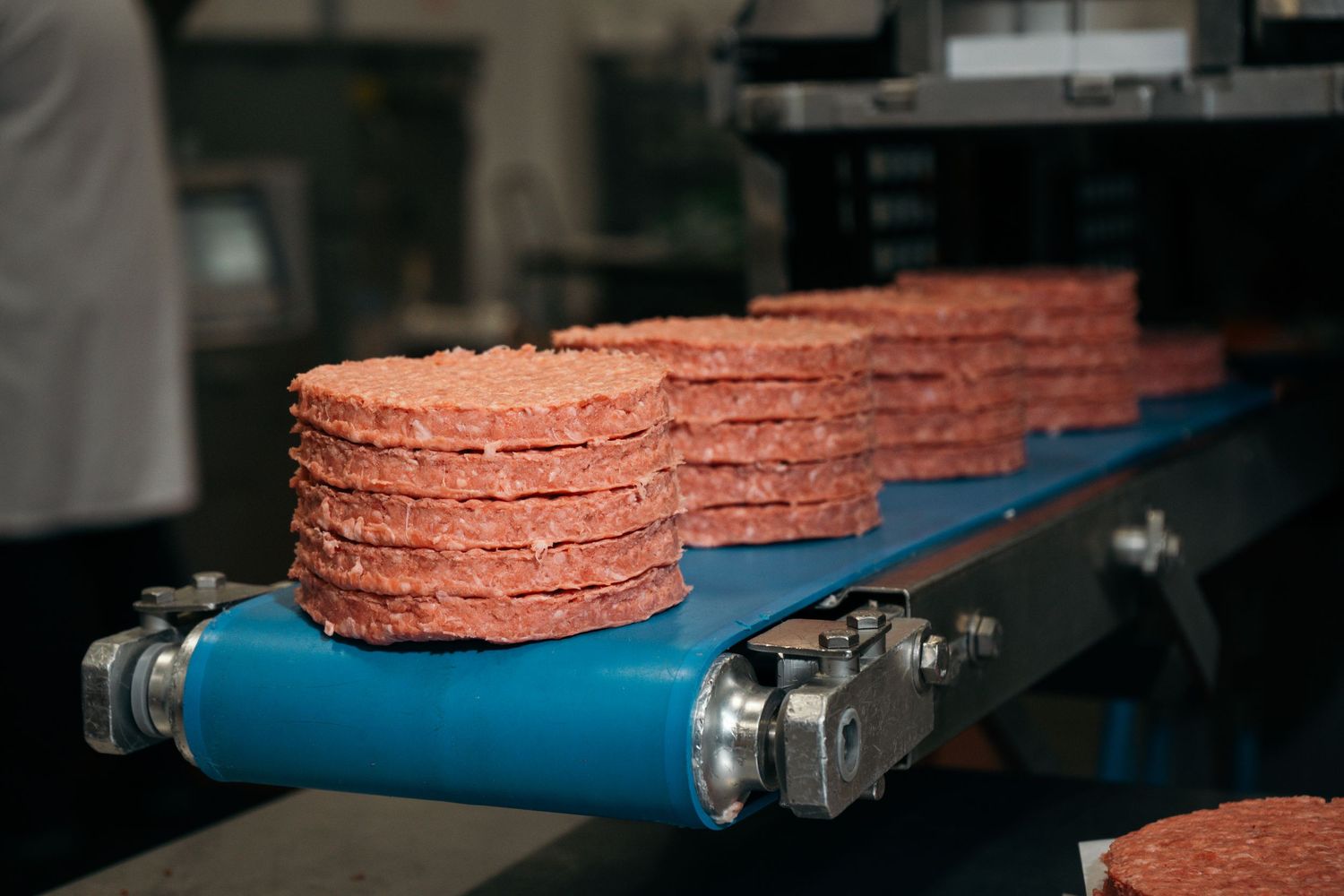 Stacks of raw beef burger patties on a blue industrial conveyor belt in a meat processing facility.