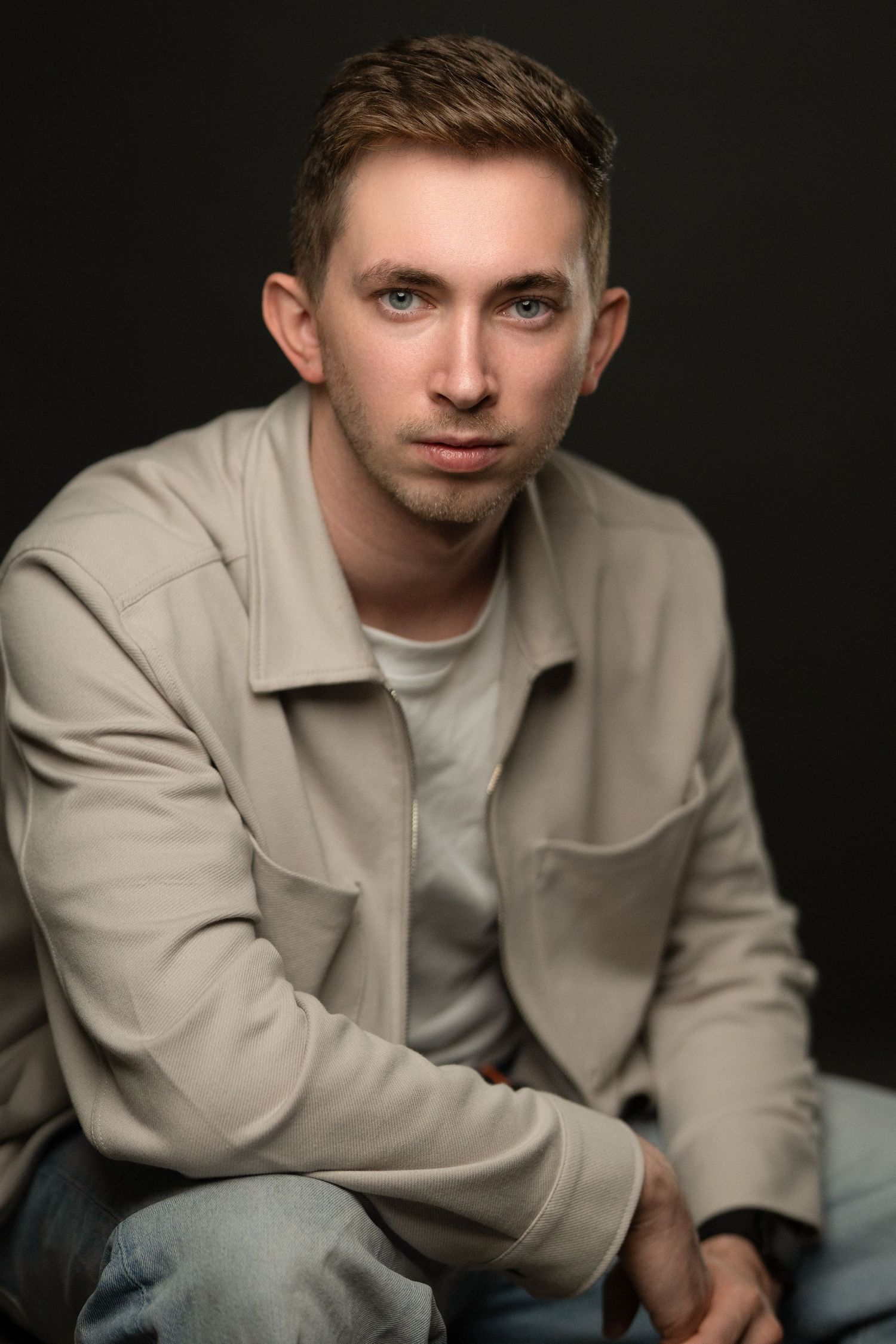 Young man with blue eyes in beige jacket poses confidently against dark studio background.