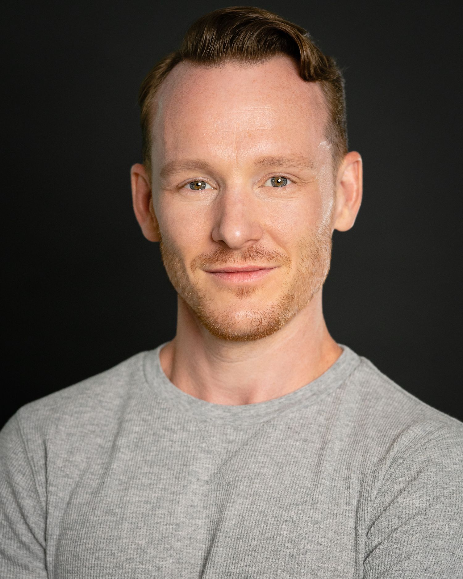 Professional headshot of a smiling red-haired man in a gray t-shirt against a dark background.