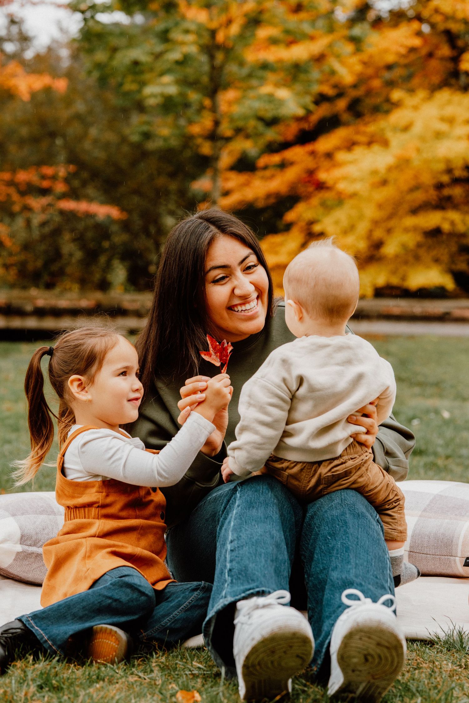 A smiling mother sits outdoors in autumn park holding baby while young daughter shares a red leaf.
