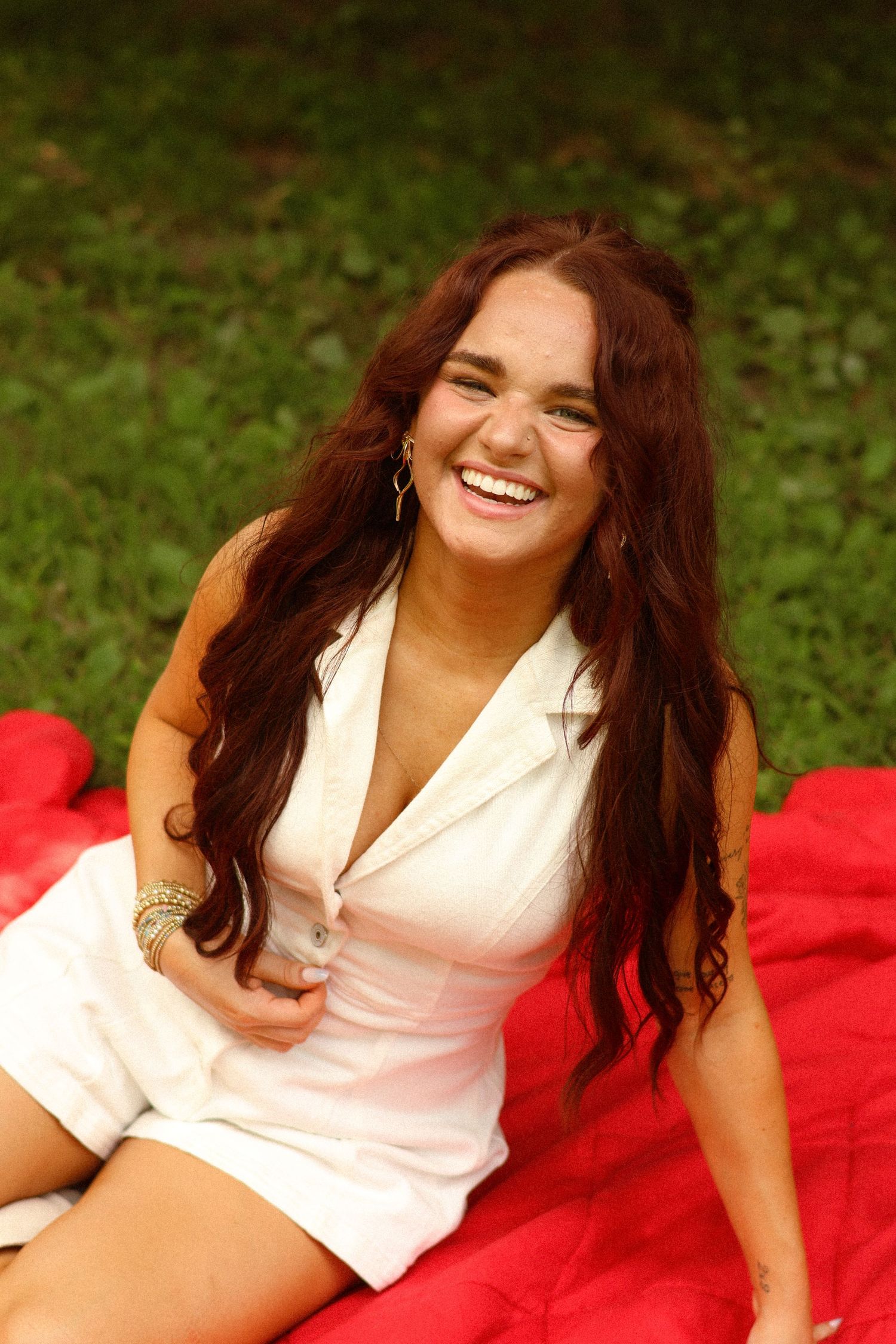 Young woman with long auburn hair laughing joyfully while sitting on a red blanket outdoors in a white dress.