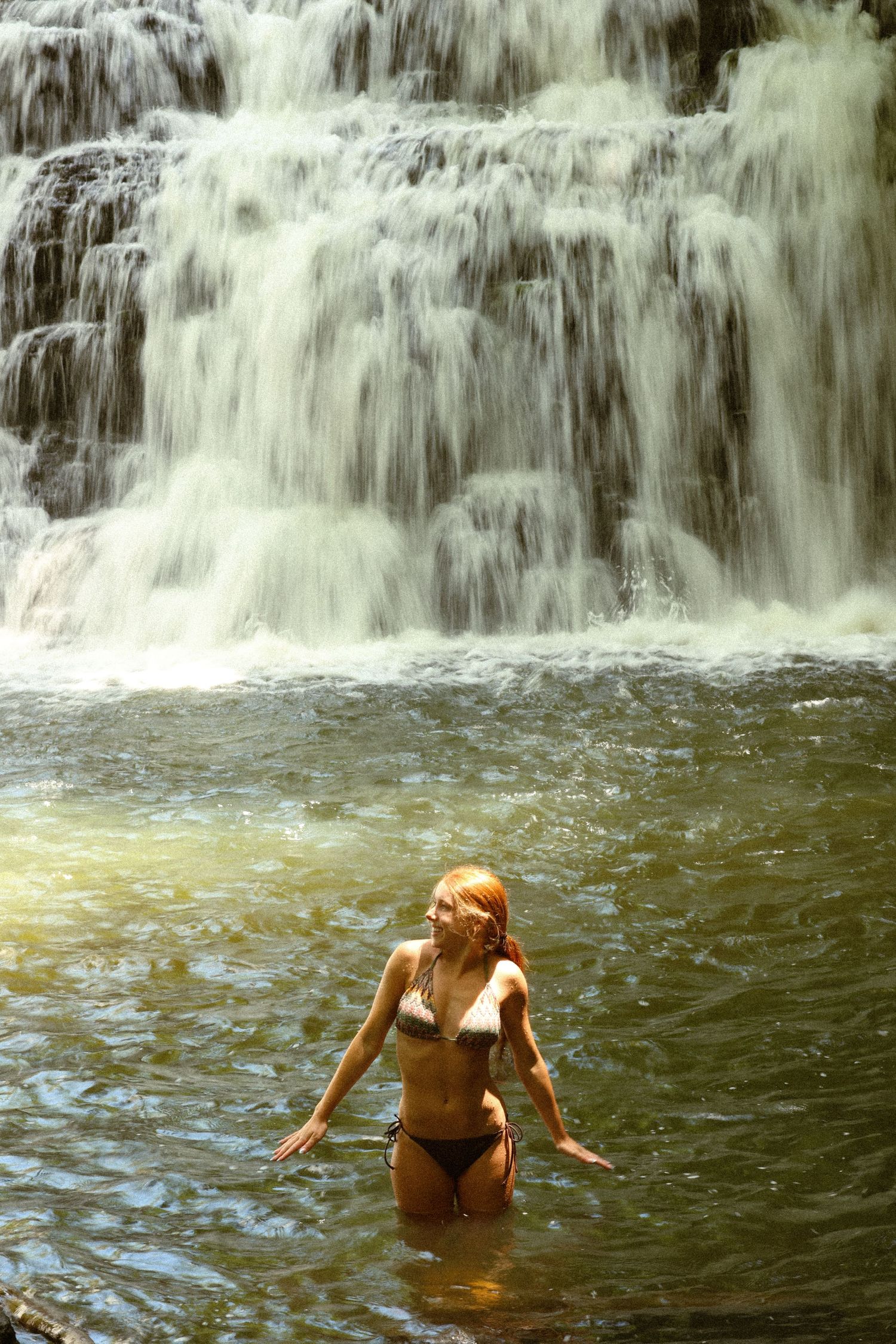 Red-haired woman in white bikini standing in a natural pool before a stunning multi-tiered waterfall.