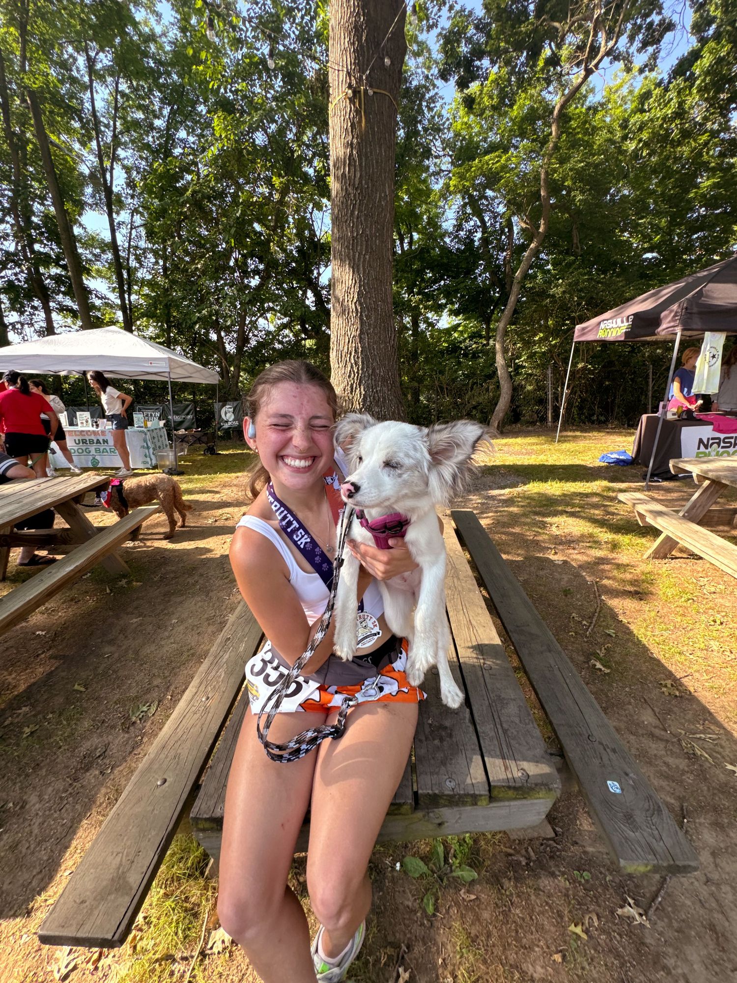 Smiling woman holding a white fluffy dog at an outdoor picnic area with trees and event tents in the background.