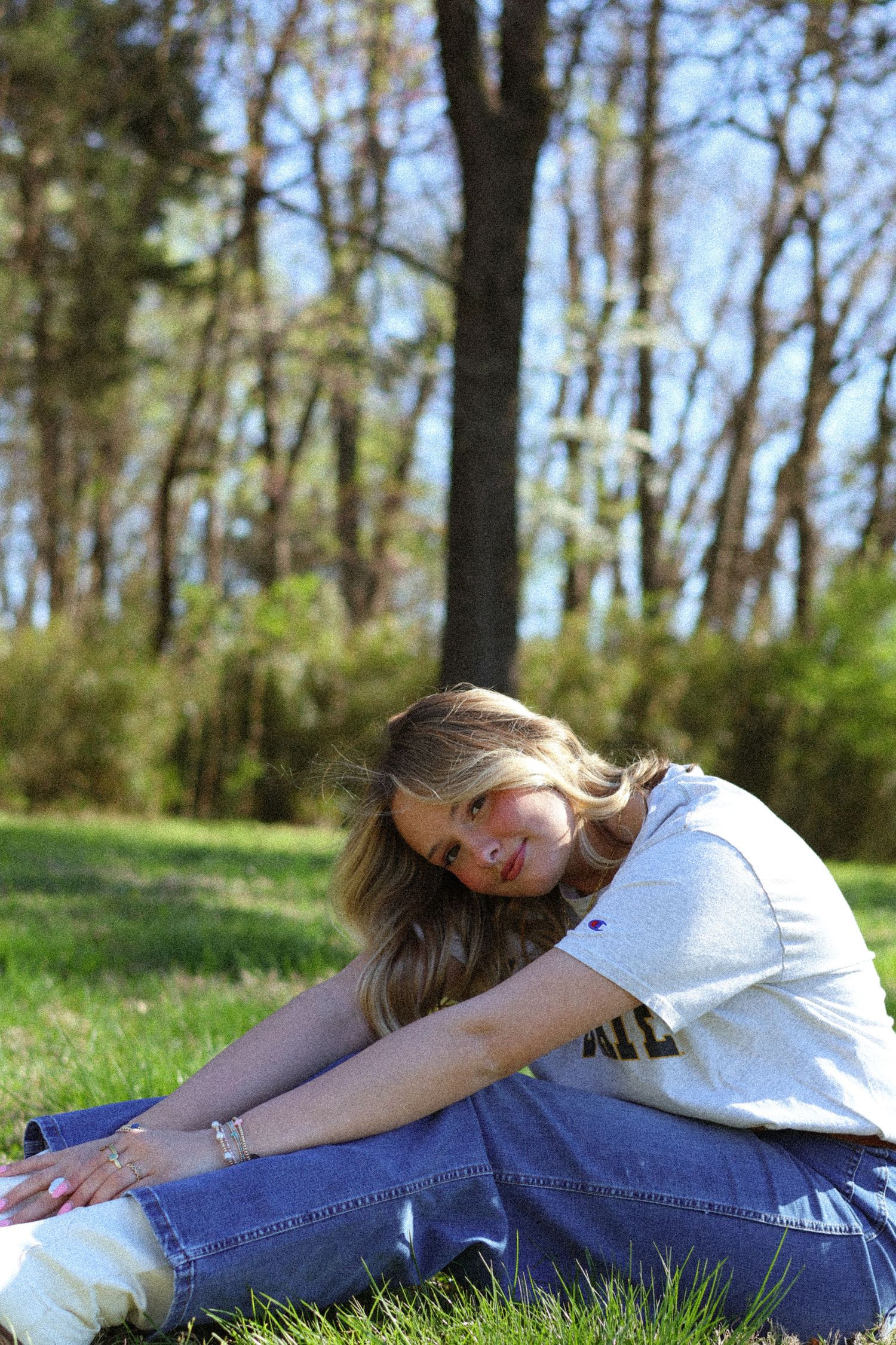 Young woman with long blonde hair sits on green grass in a sunny park, wearing jeans and a gray t-shirt.