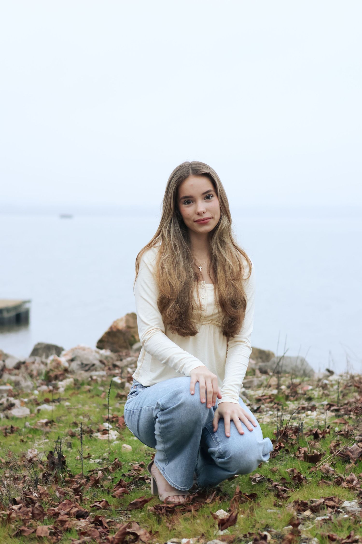 Person in white sweater and jeans sits casually on ground covered with fallen autumn leaves.