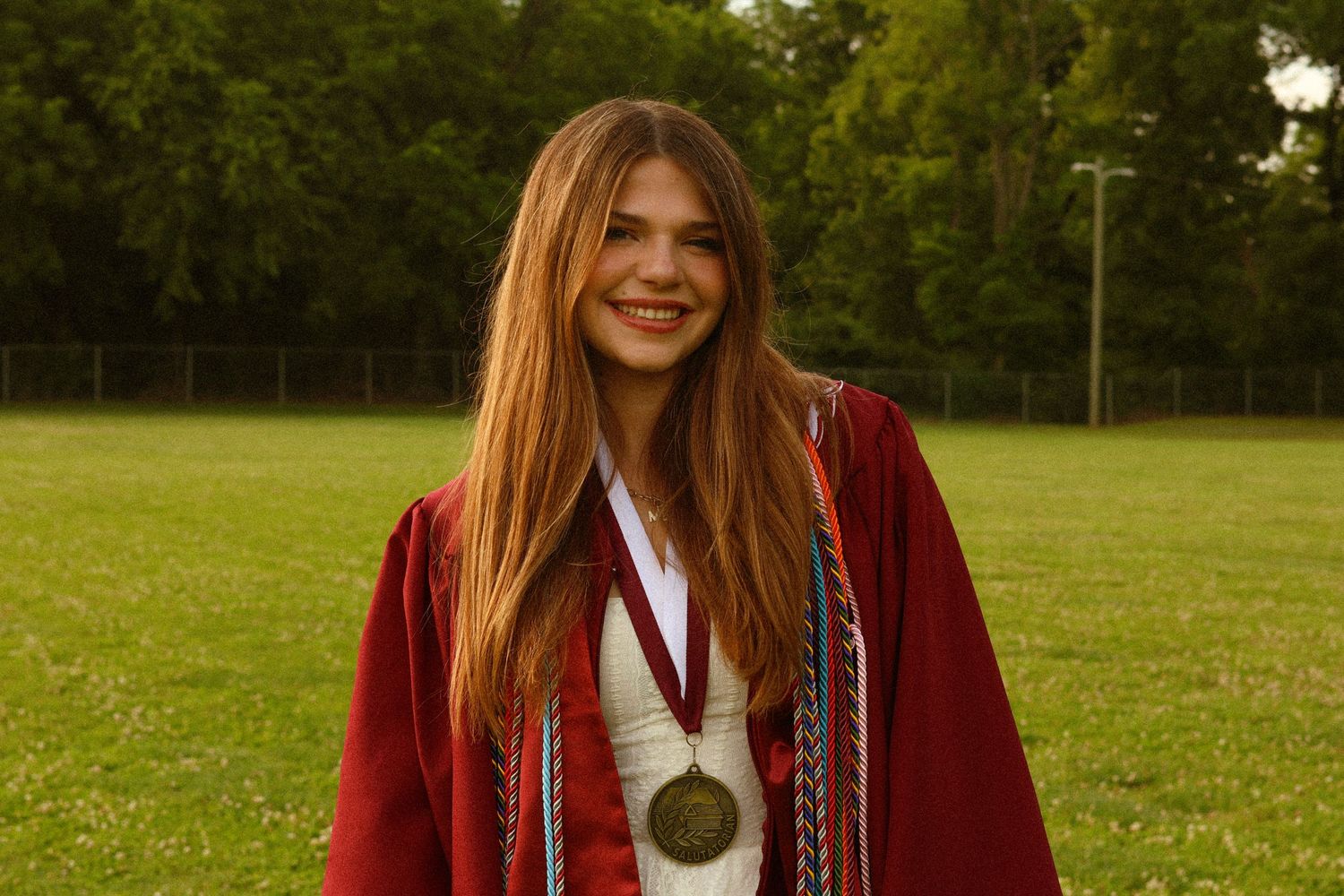 Smiling young woman in red graduation gown with medal and honor cords standing on green field outdoors.