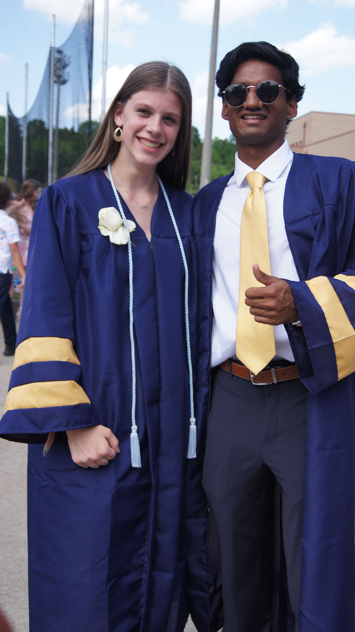 Two graduates in blue academic regalia with gold accents pose together at commencement ceremony outdoors.