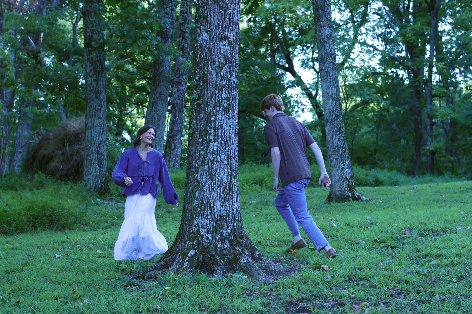 Couple playing and dancing around tree in wooded outdoor setting.