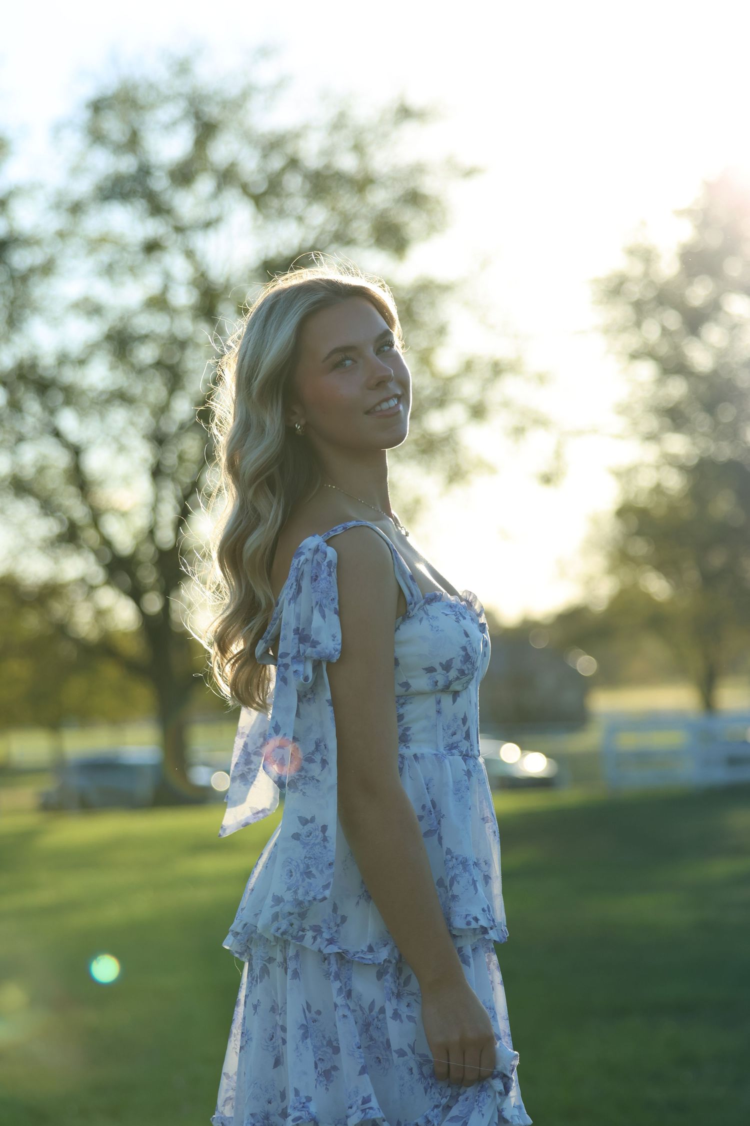 A young girl in a white floral dress poses outdoors at sunset in a grassy field with backlit sunlight creating a golden glow.