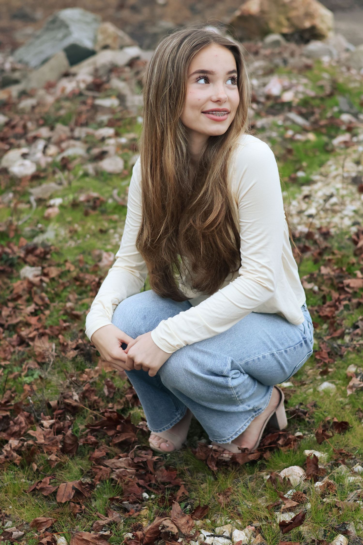 Person in casual white sweater and denim poses on ground with scattered fall foliage.