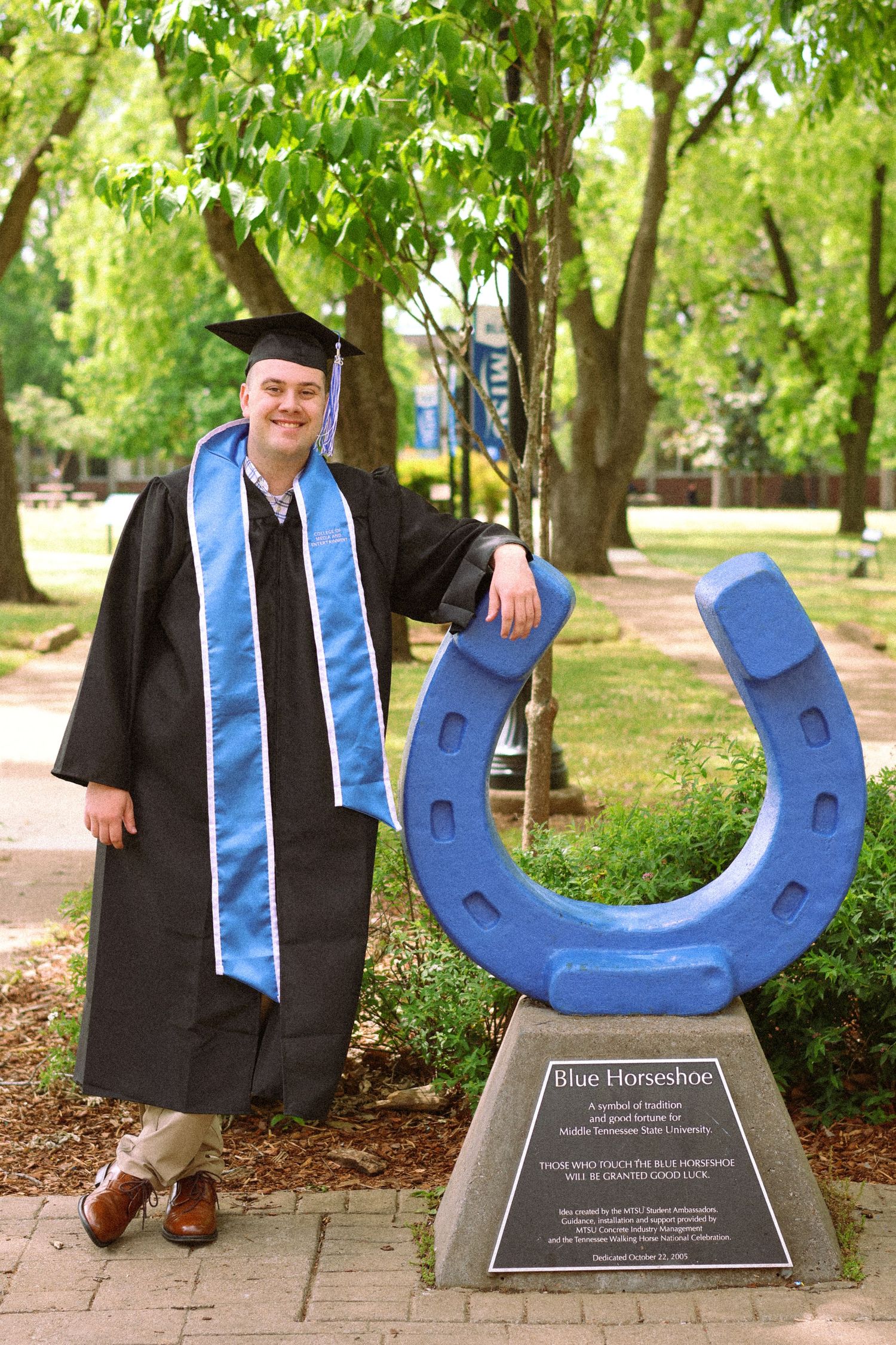 Graduate in cap and gown poses beside a blue horseshoe sculpture on a tree-lined campus walkway.