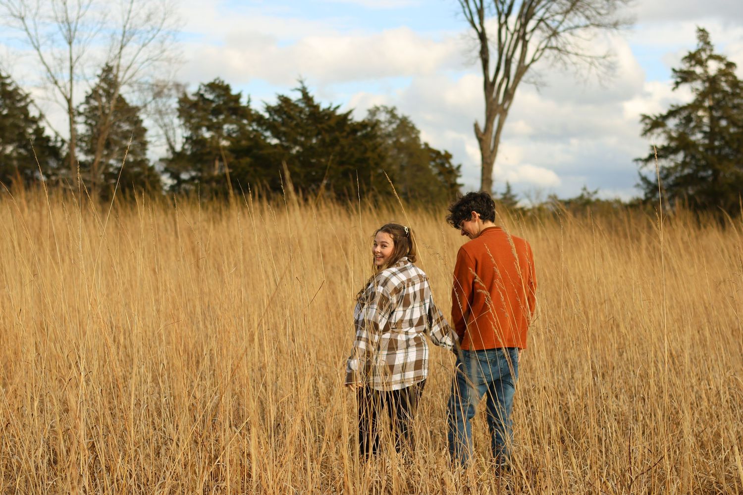 Two people stand together in a golden autumn field of tall grass with bare trees in the background.