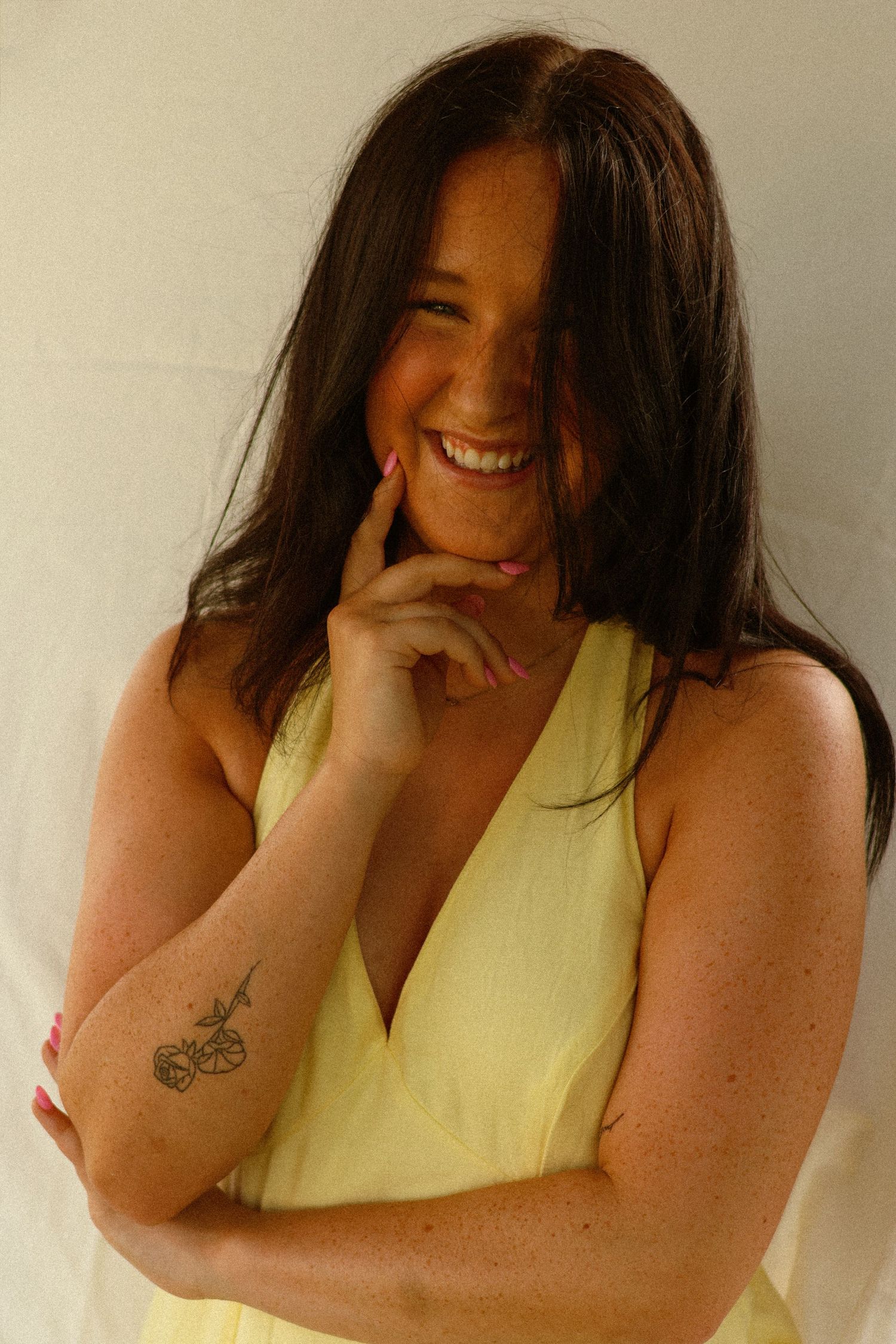 Laughing brunette woman in yellow dress showing tattoos while posing playfully with hand on chin.