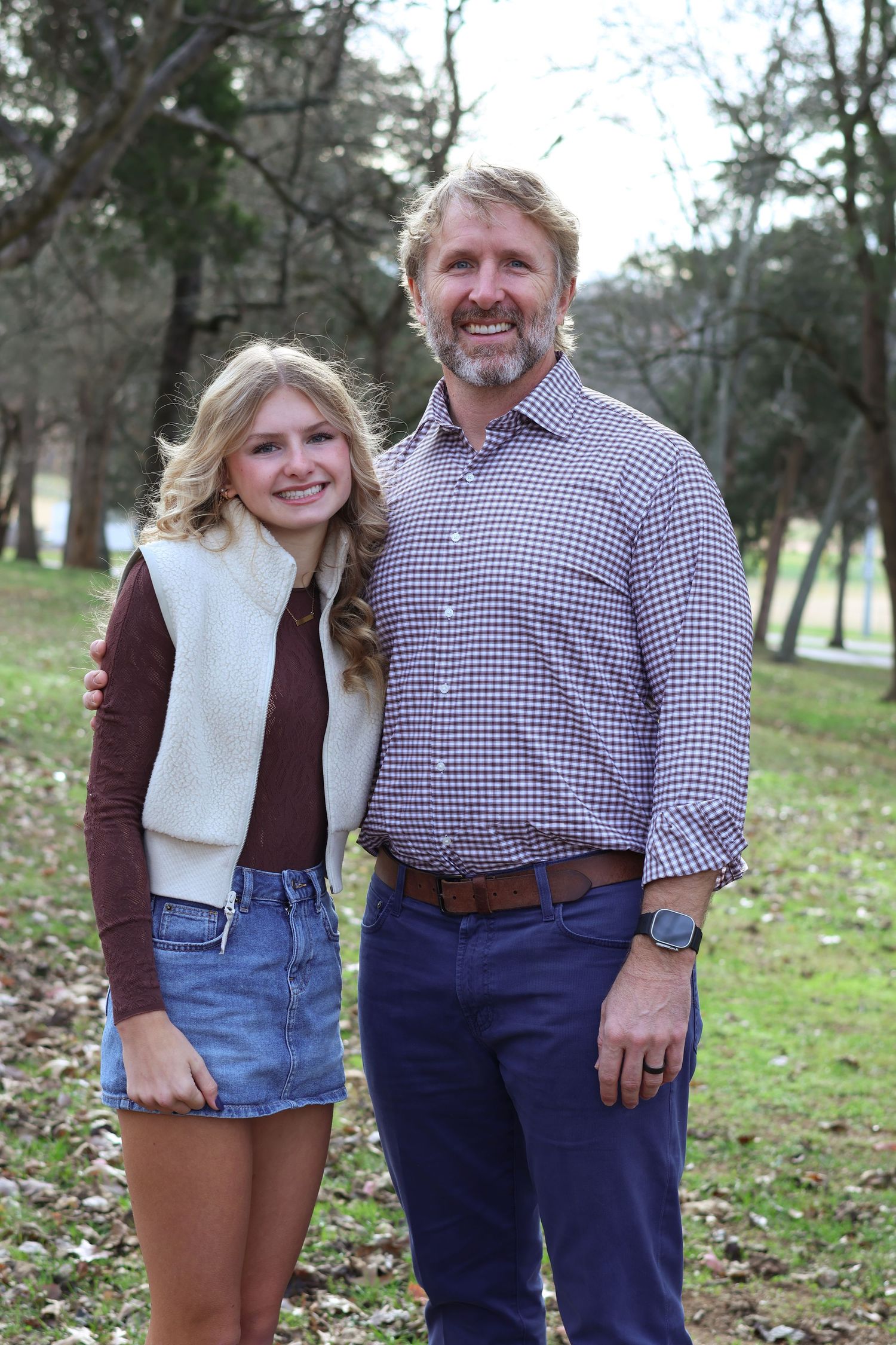 A parent and child stand together outdoors in casual fall attire with trees in the background.