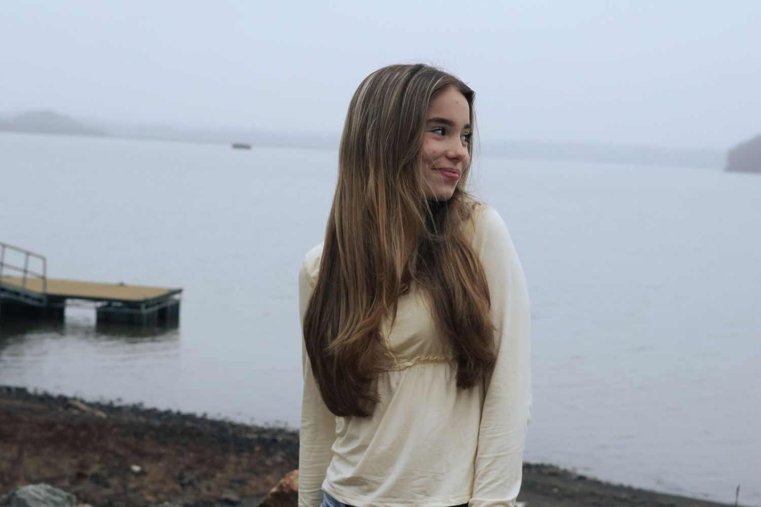A person in a cream sweater stands near a misty lake on an overcast day.