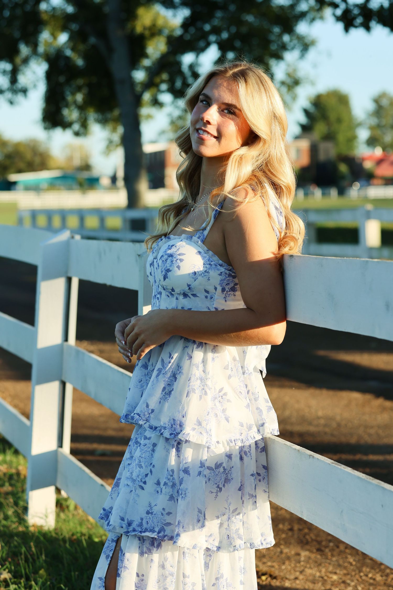 A child in a white floral dress plays near a white picket fence on a sunny afternoon.