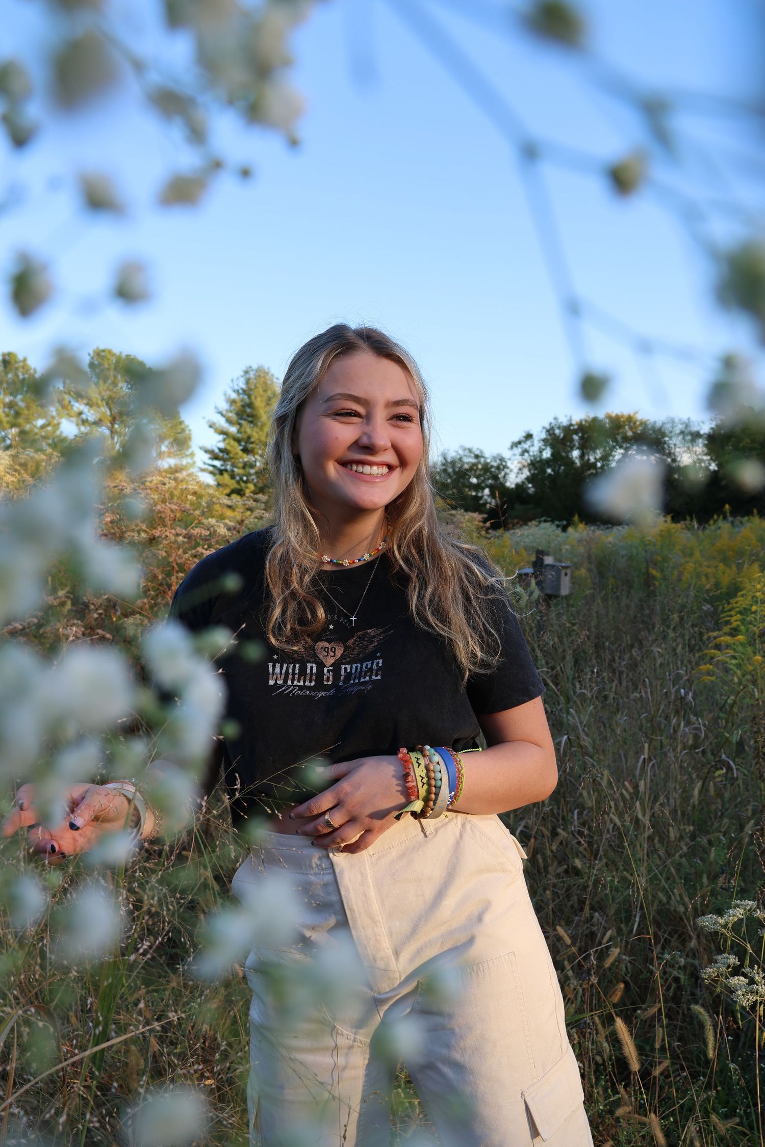 A person in a black t-shirt and white pants poses playfully in a sunny wildflower field during golden hour.