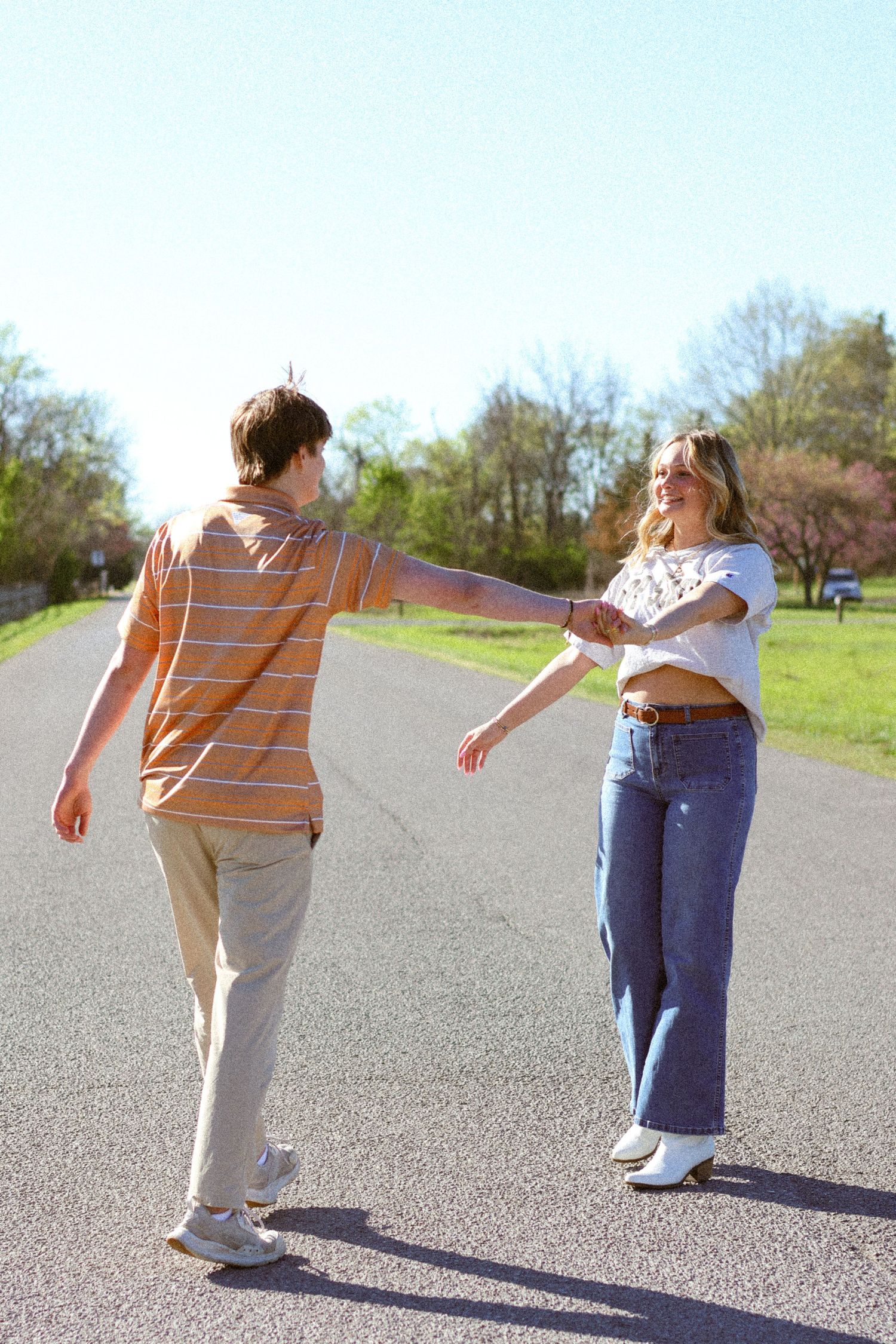 A young couple holding hands on a sunny road, the woman smiling in jeans and white top, man in striped shirt.