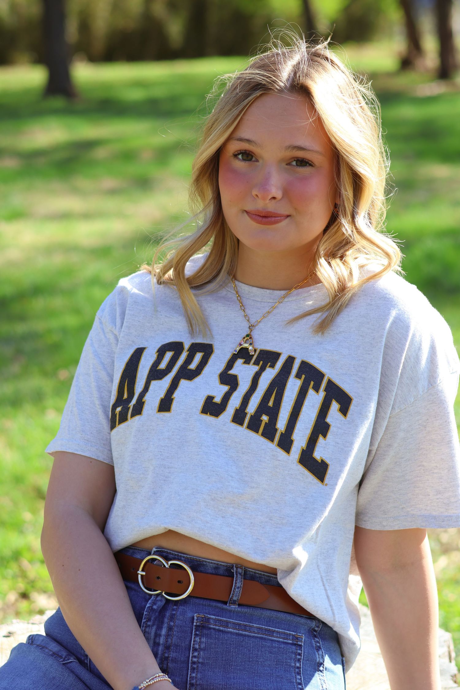 Young woman wearing an App State gray graphic t-shirt and jeans, posing outdoors in a sunny park setting.