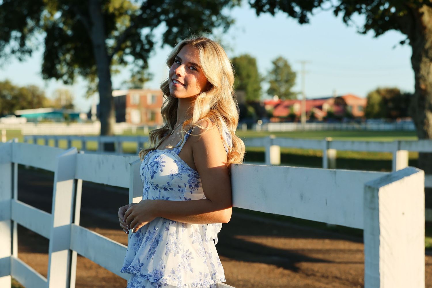 Woman in white lace dress smiles while leaning against white fence on sunny day near water.