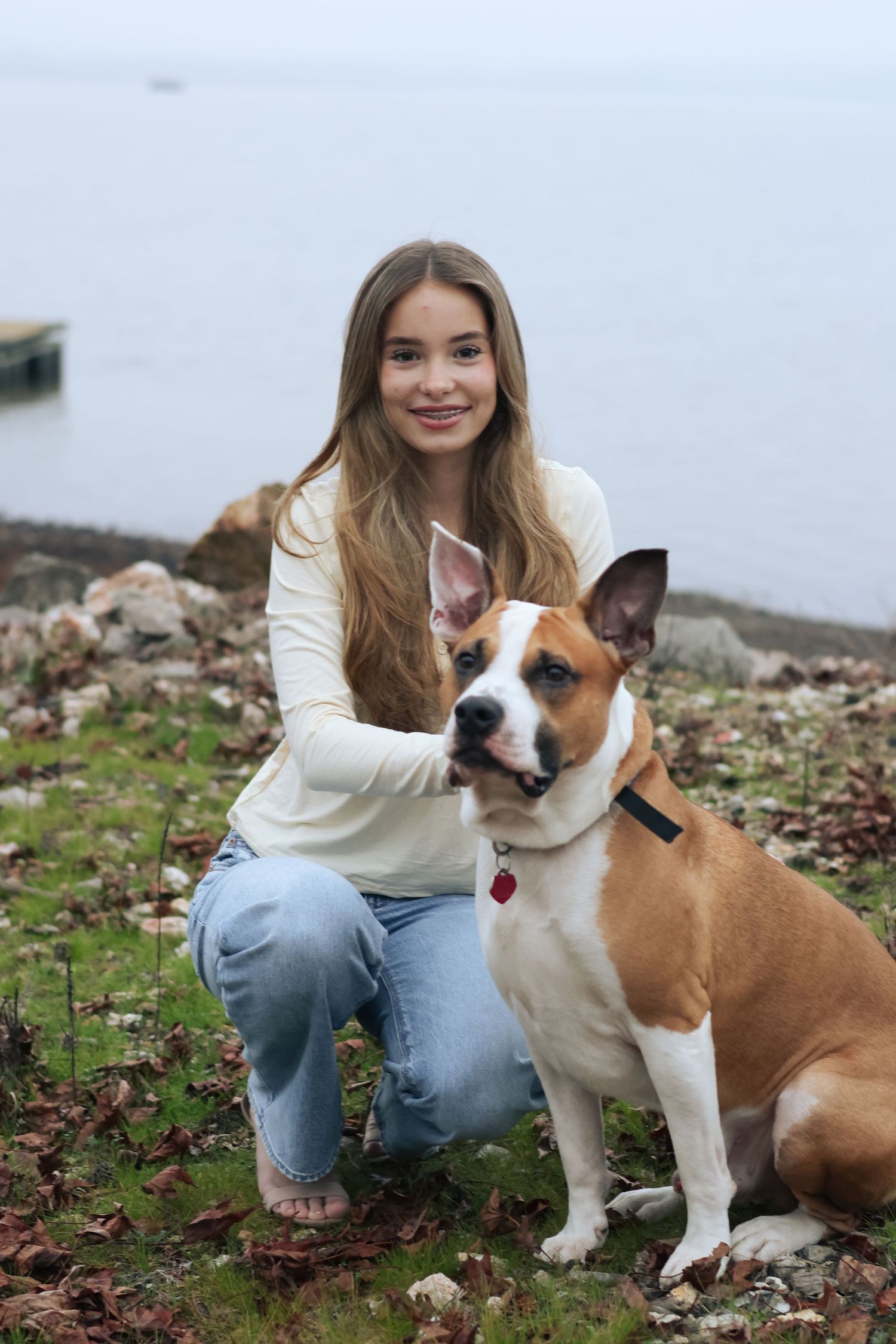 A person in jeans and white sweater sits on the ground with two dogs, one brown and white and one brown.