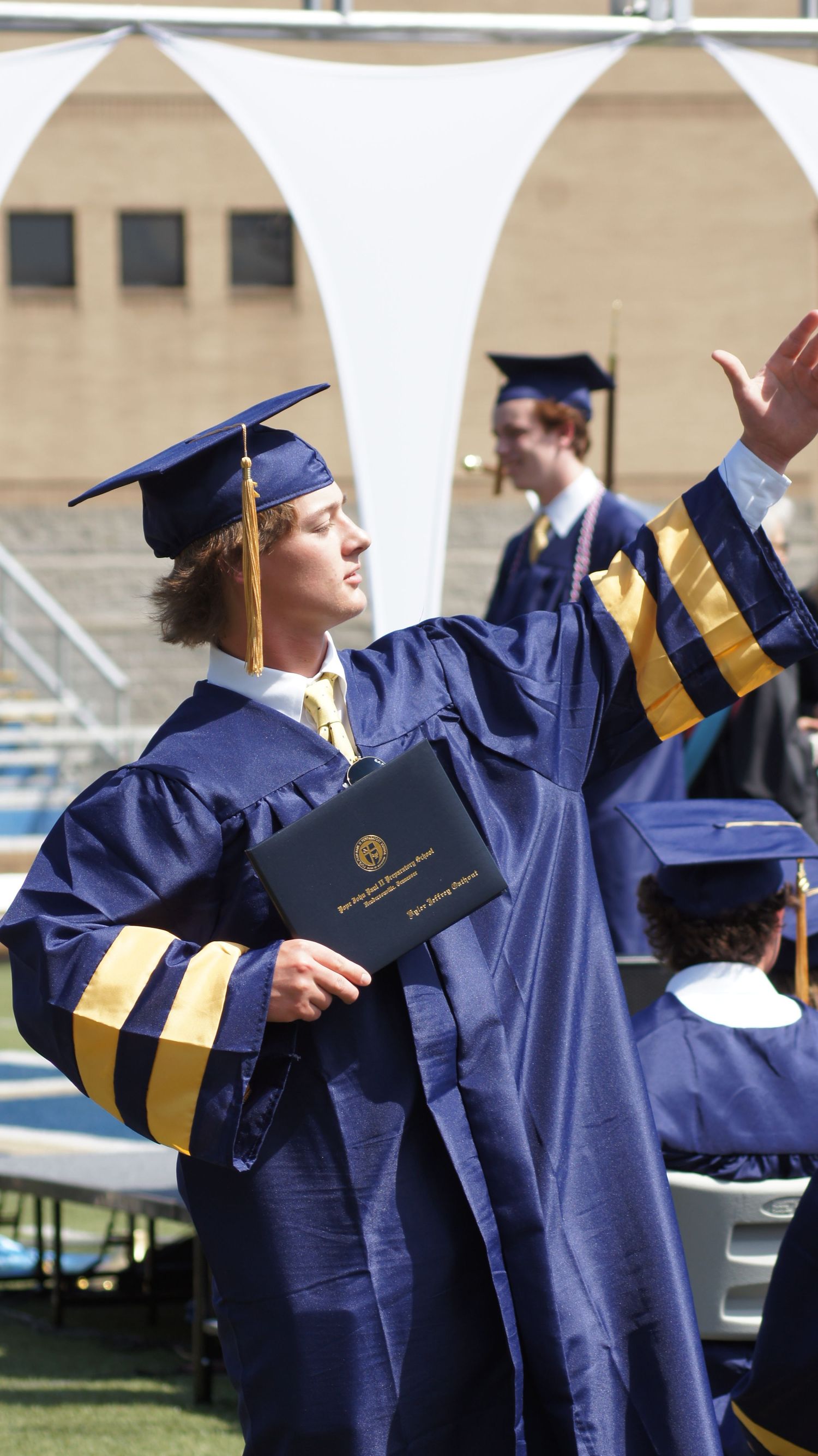 A graduate in navy blue and gold regalia celebrates at commencement while holding their diploma and raising one arm.