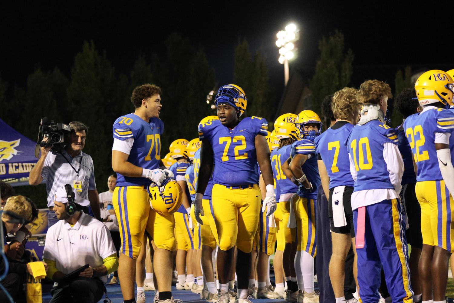 Football players in blue and yellow uniforms gather on the sideline during a night game.