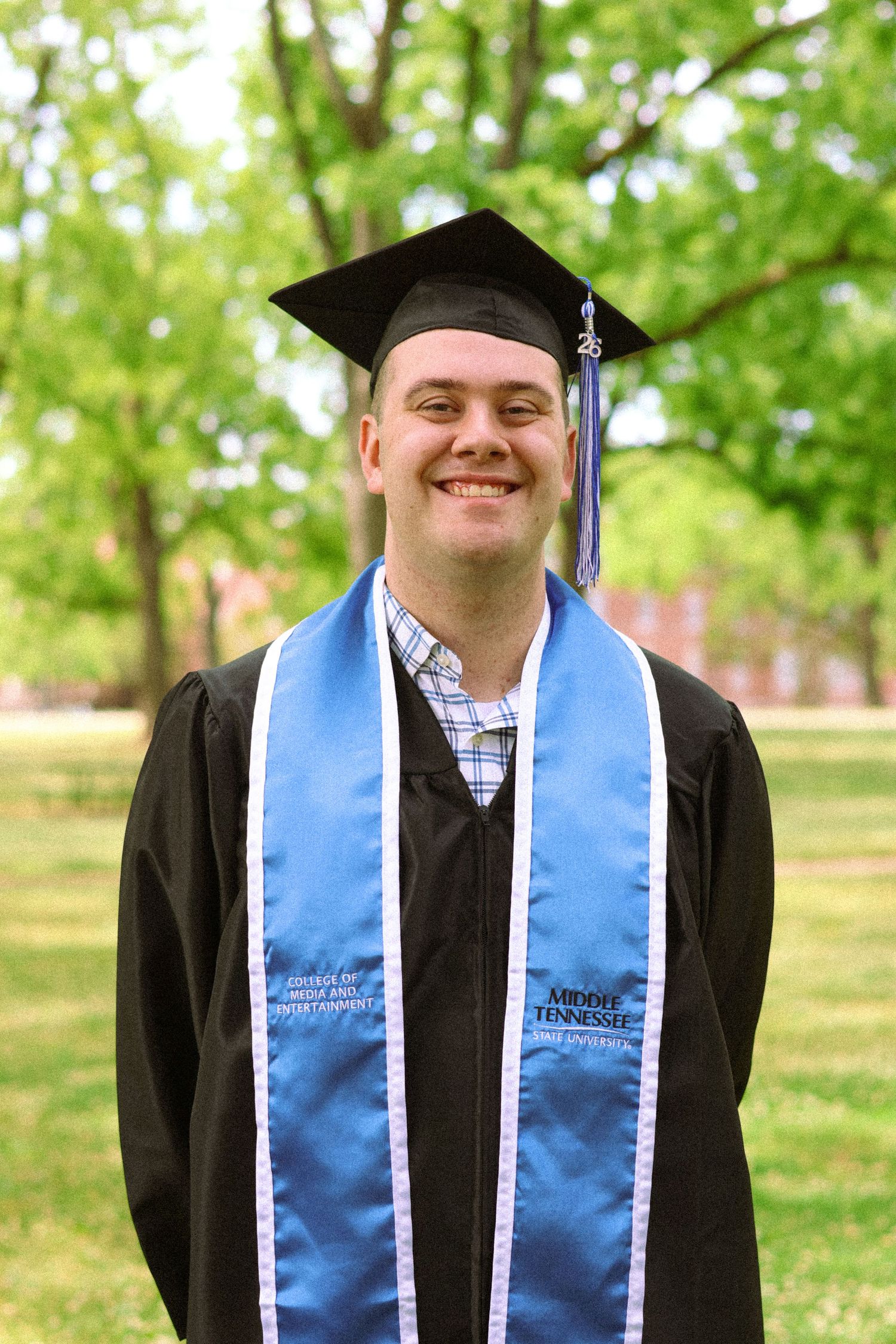Smiling graduate in black cap and gown with Middle Tennessee blue stole poses outdoors among green trees.