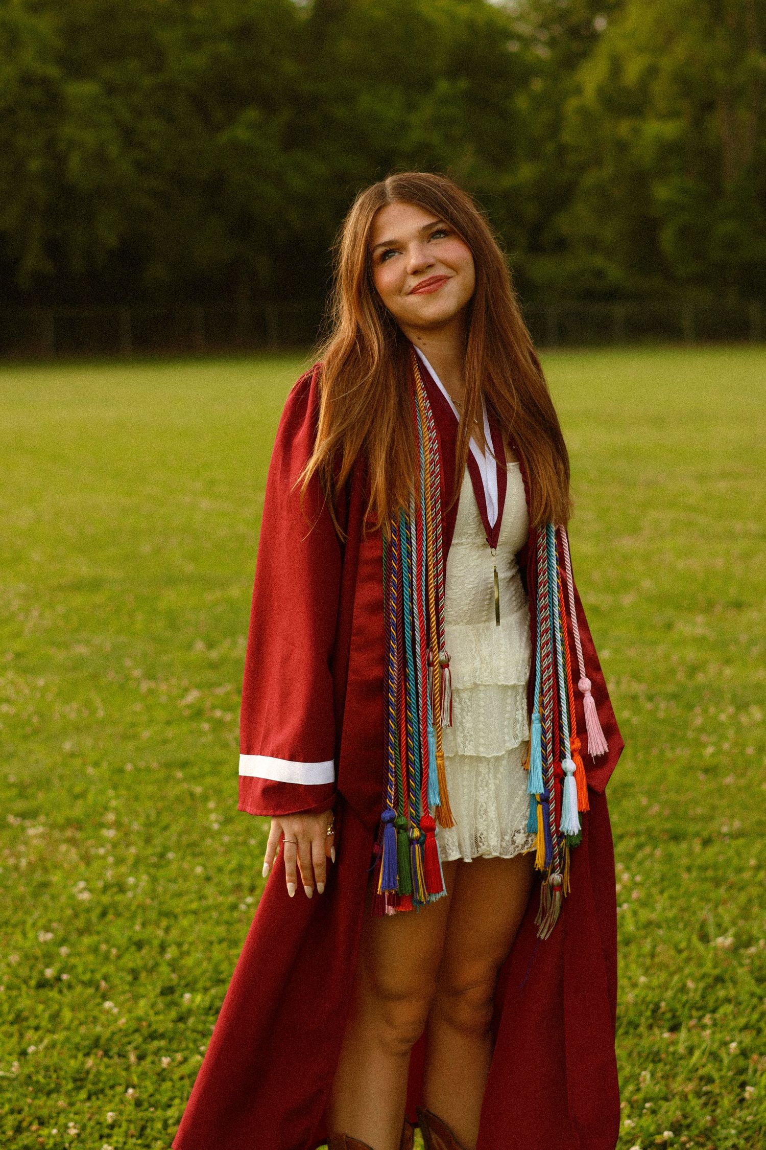 Young woman in red graduation gown with colorful honor cords smiles outdoors on green grass field.