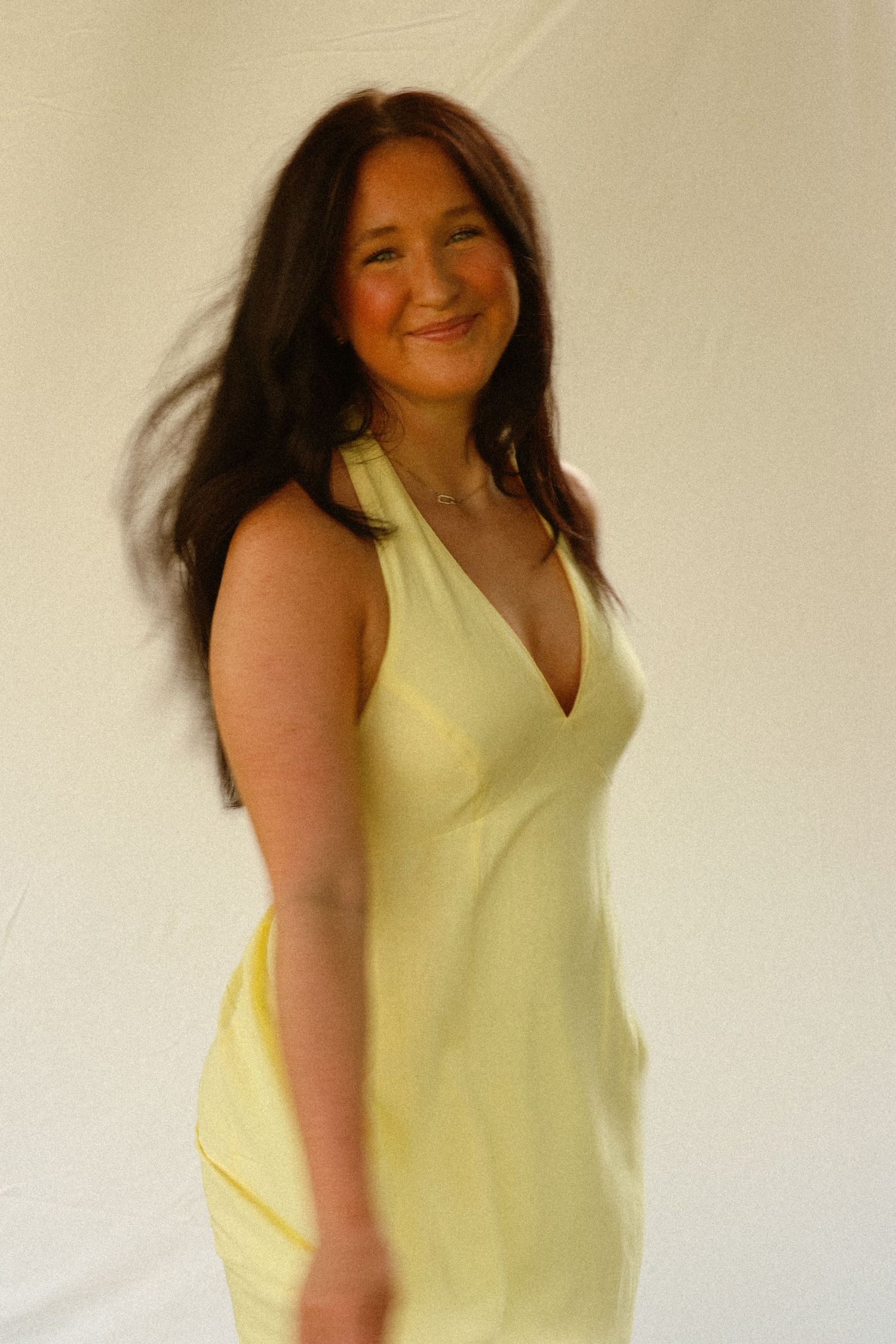 Smiling woman with dark hair wearing a yellow sleeveless v-neck dress, posing against a white background.