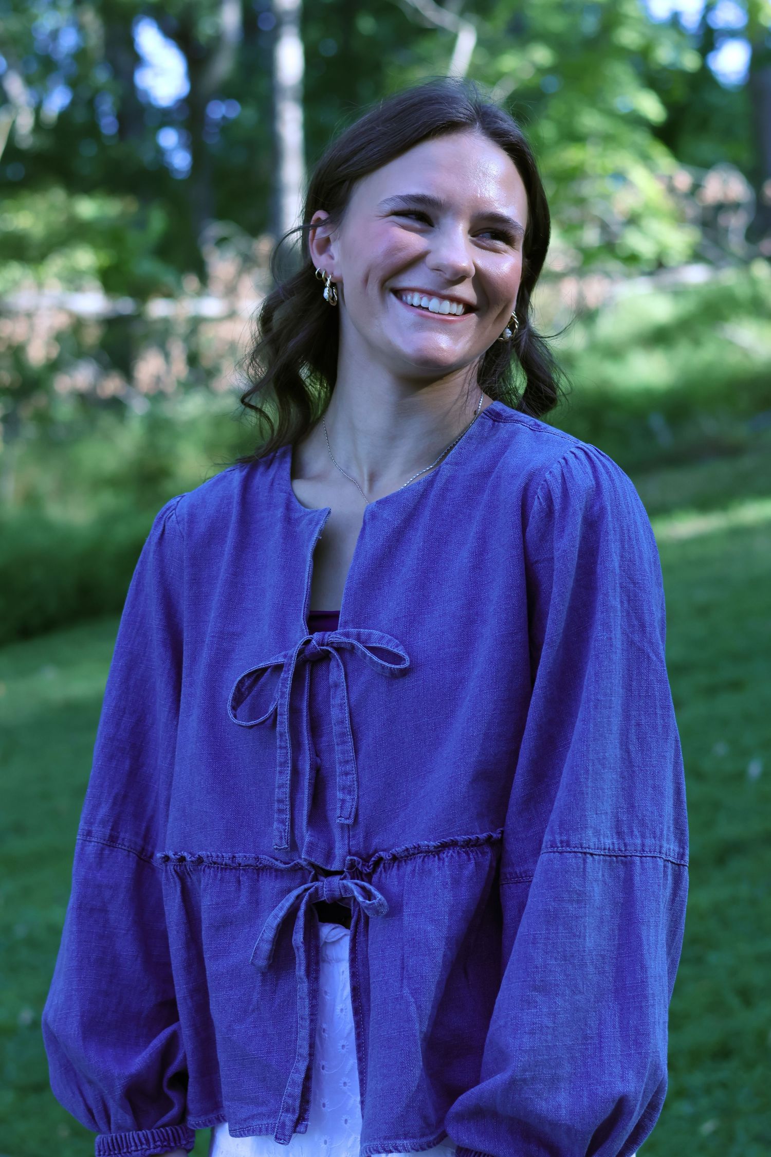 Woman in purple peasant blouse smiles brightly in outdoor garden setting.