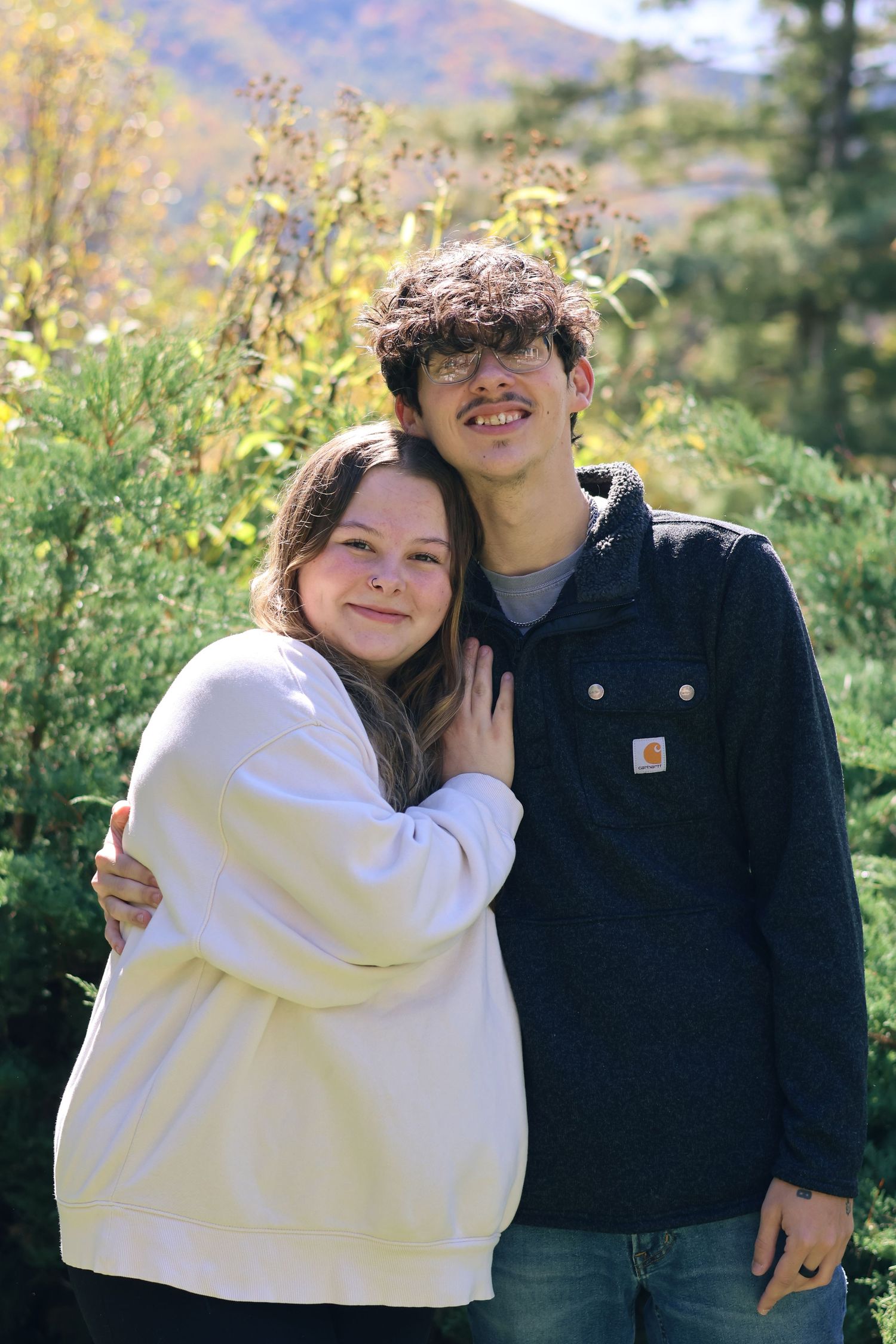A happy couple in matching light and dark outfits embraces outdoors against a mountain backdrop.