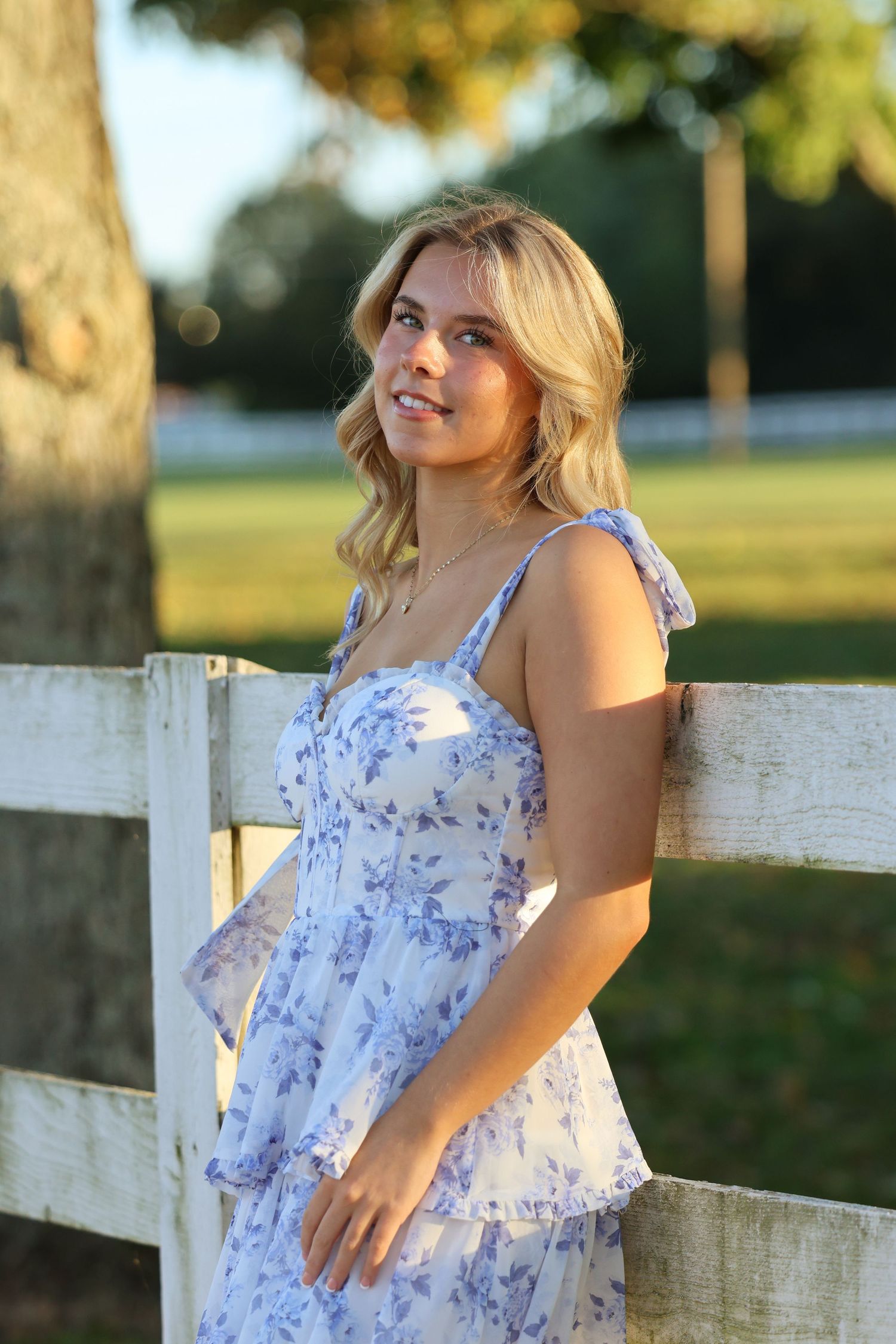 A person in a blue and white floral sundress poses by a white wooden fence at sunset in a park setting.