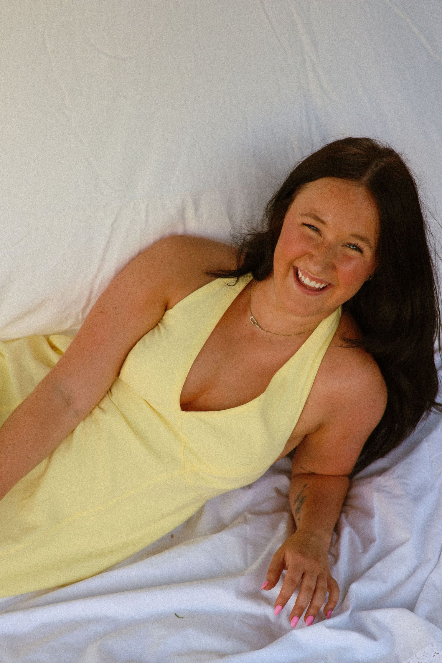Happy woman with dark hair in a yellow halter dress lying down laughing against a white background.