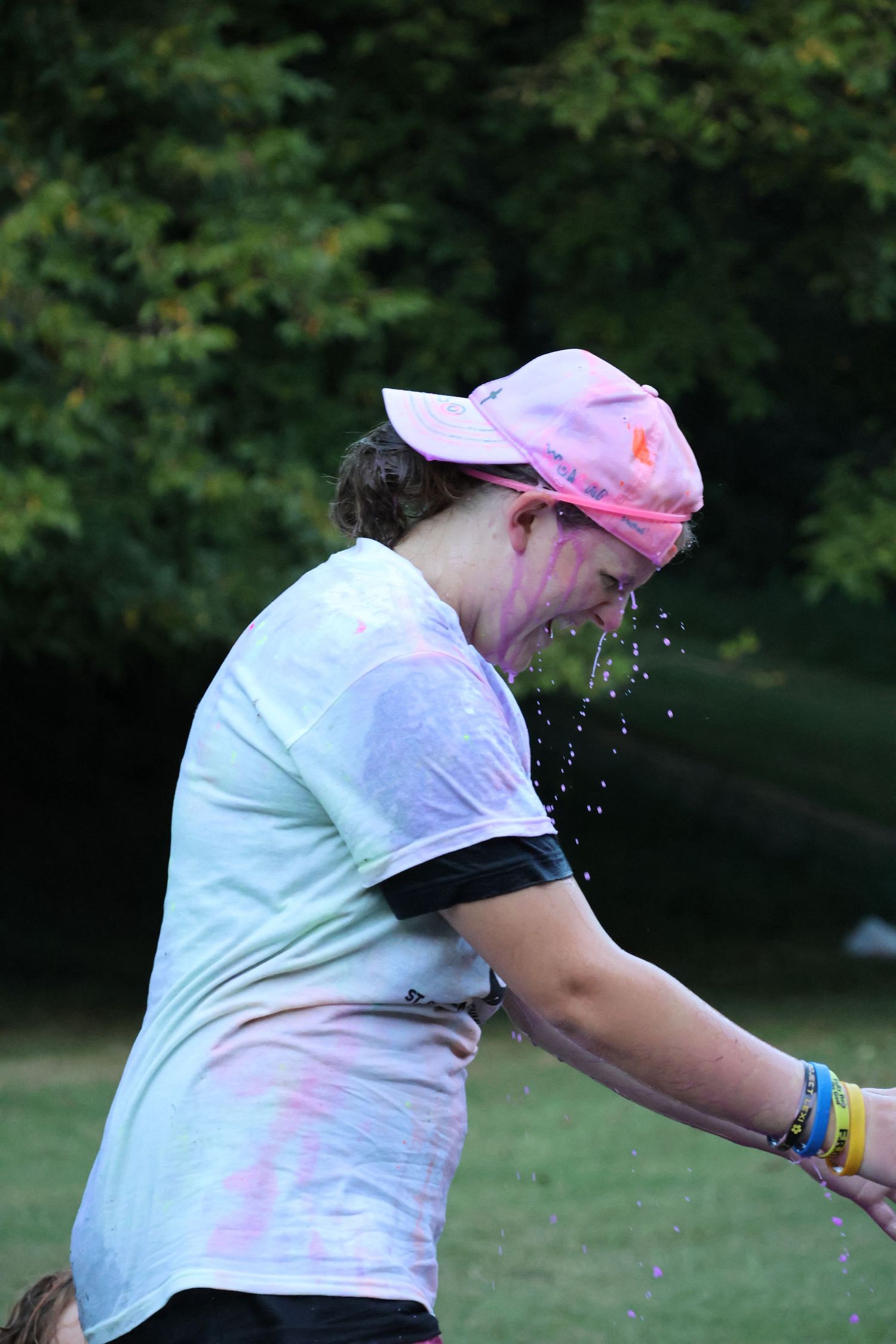 Person in white t-shirt and pink cap splashing colorful powder during outdoor color run event.