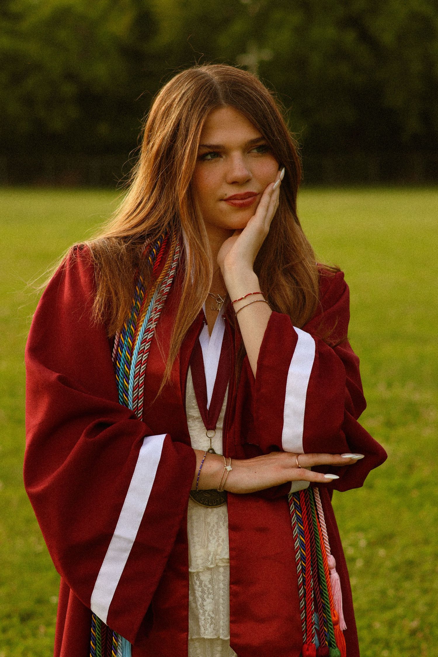 Young woman in red graduation gown with colorful stoles poses thoughtfully outdoors on green grass.
