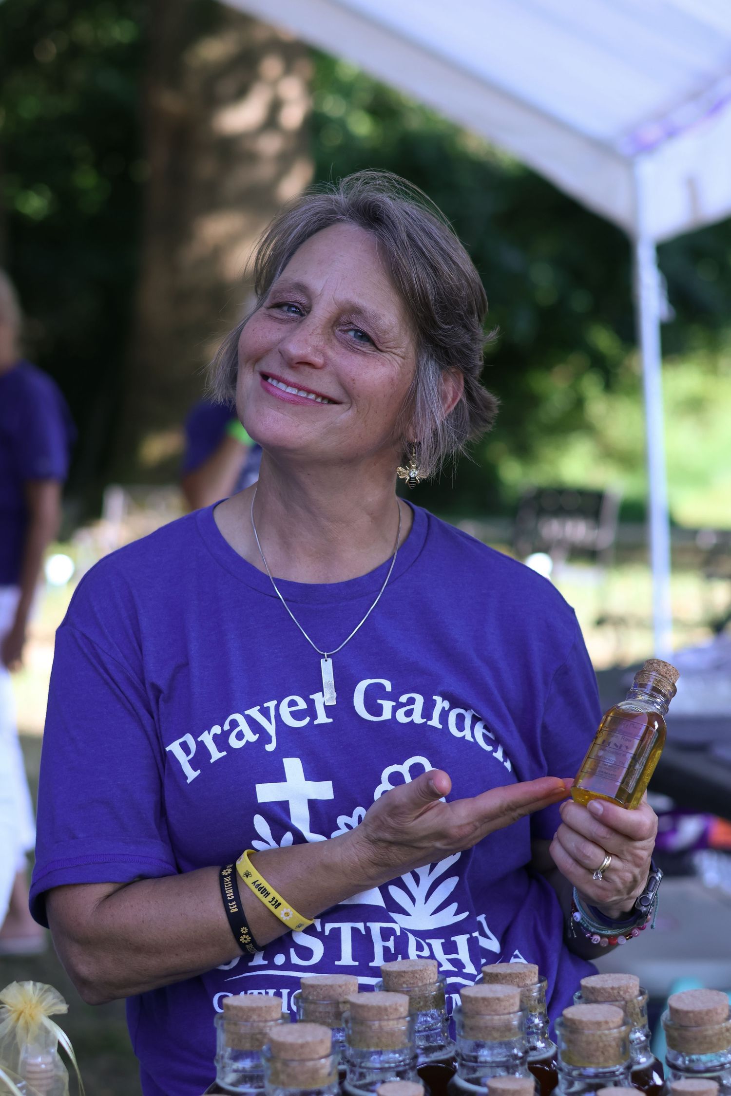 Person in purple Prayer Garden t-shirt smiles while handling items at outdoor event.