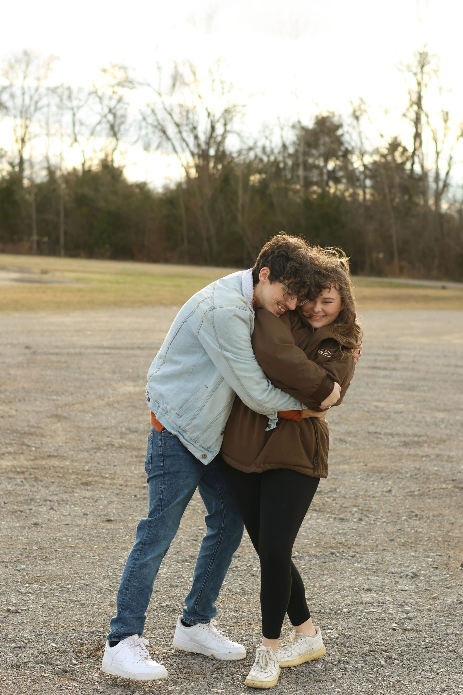 A couple in casual denim and brown attire share an intimate moment in an open field at sunset.