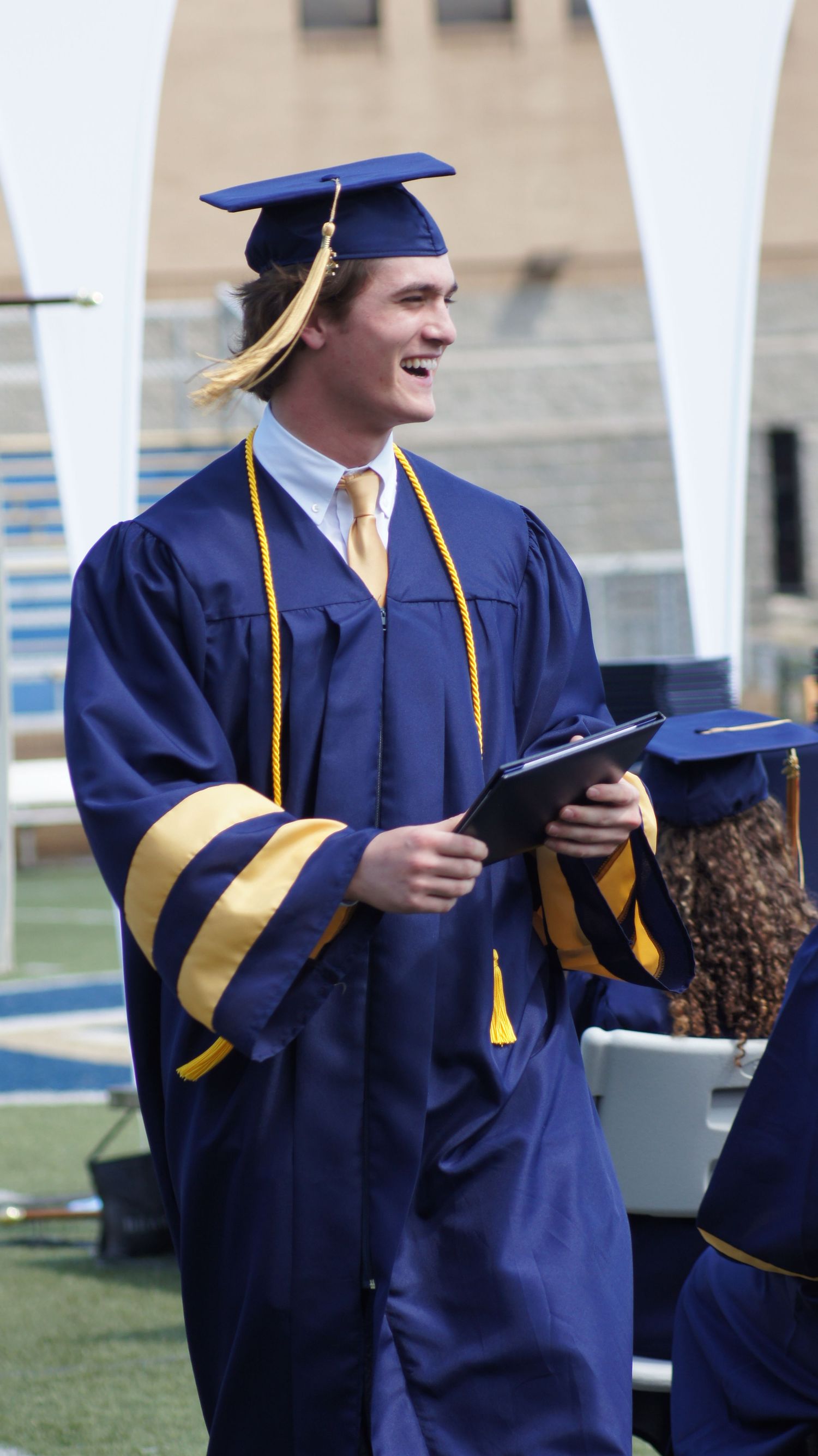 Graduates in navy blue caps and gowns with gold trim celebrate during commencement ceremony outdoors.